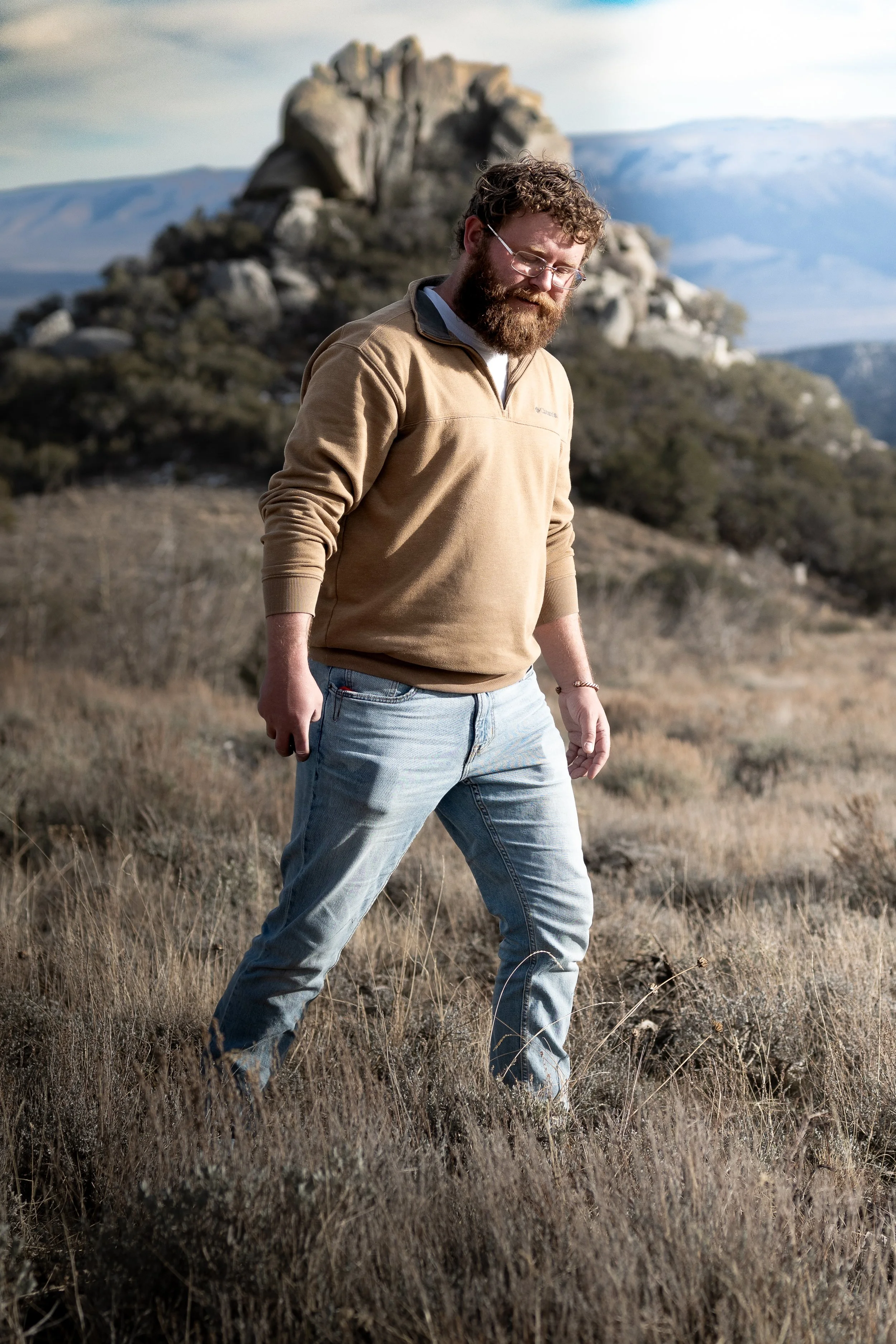 A man with glasses and a beard walking outdoors in a grassy, rocky landscape with mountains and a large rock formation in the background.