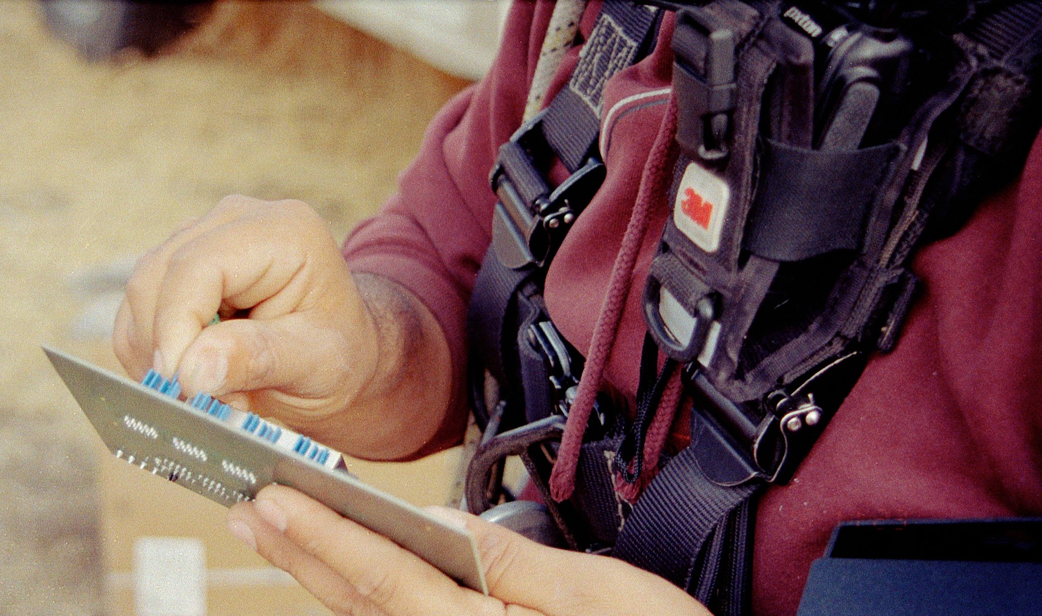 Close-up of a person's hands holding and pressing blue buttons on a timing device with a black strap, wearing a maroon shirt and a harness with various clips and tags.
