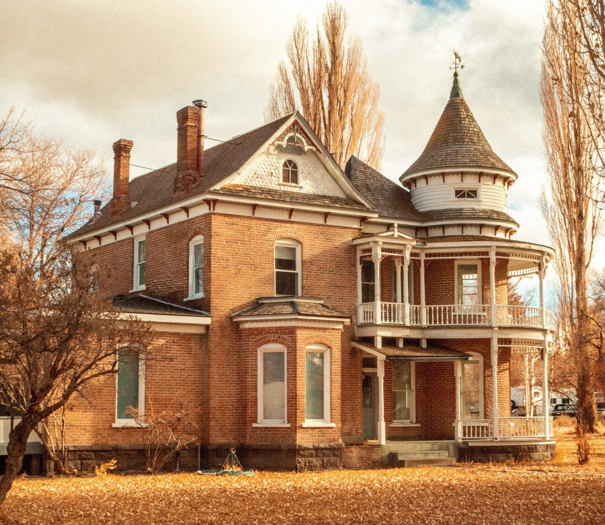 A historic Victorian-style brick house with a turret and wraparound porch, set against a backdrop of fall-colored trees and a cloudy sky.