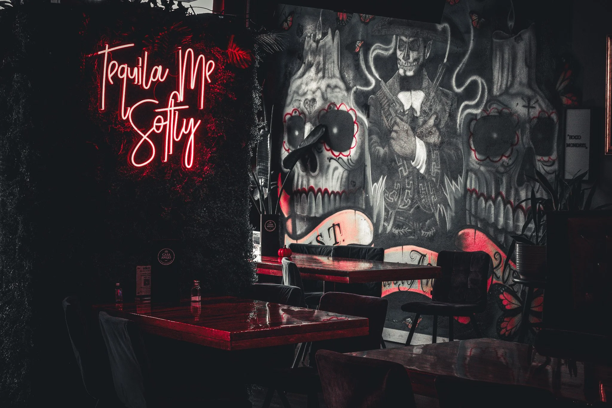 A dimly lit bar or restaurant with a neon sign that says 'Tequila Me Softly' on a wall. The wall behind has graffiti-style artwork of a skeleton dressed like a mariachi musician, surrounded by various decorative elements.