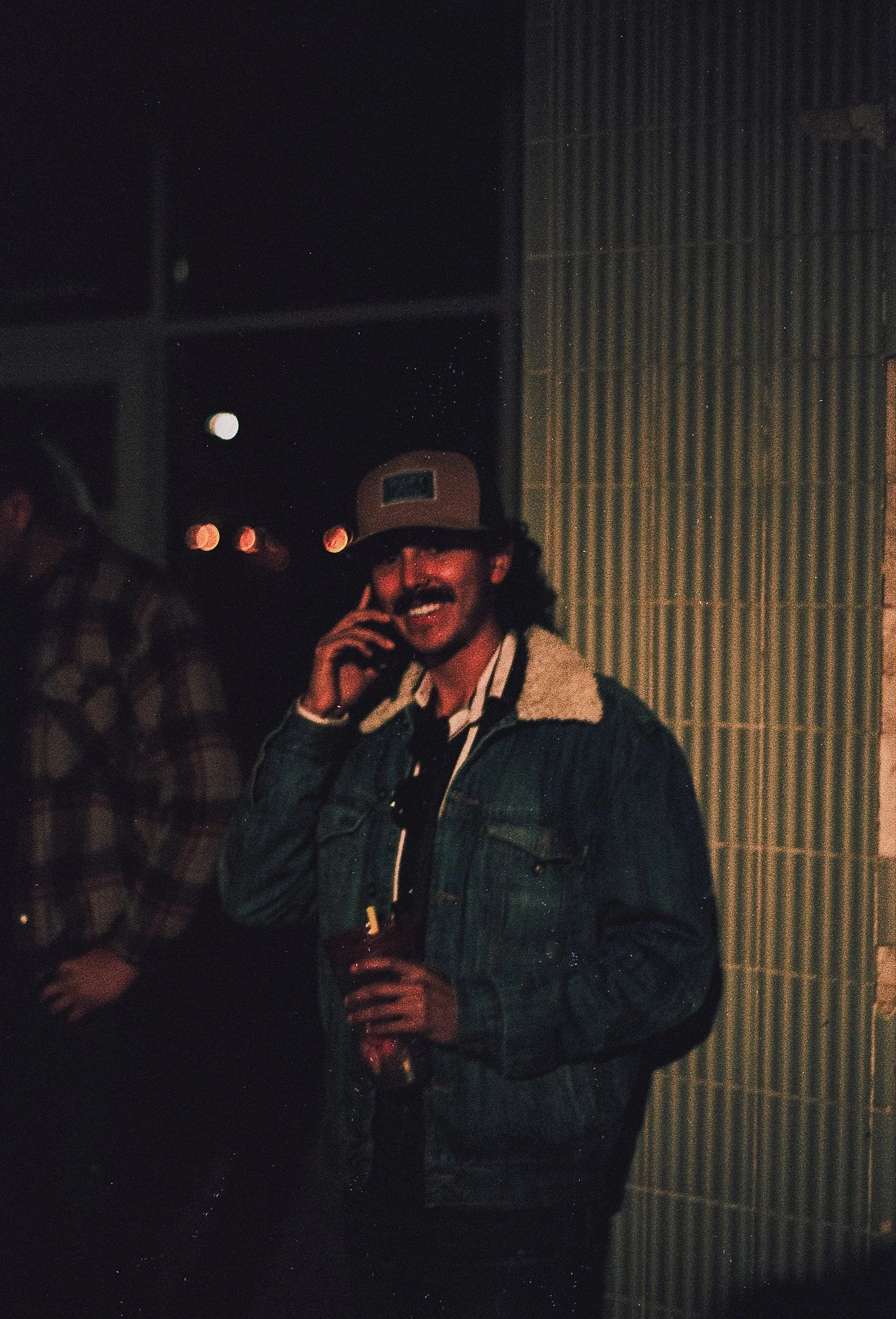 A man with curly hair, wearing a cap, glasses, and a denim jacket, smiling and holding a drink, standing in a dimly lit indoor space.