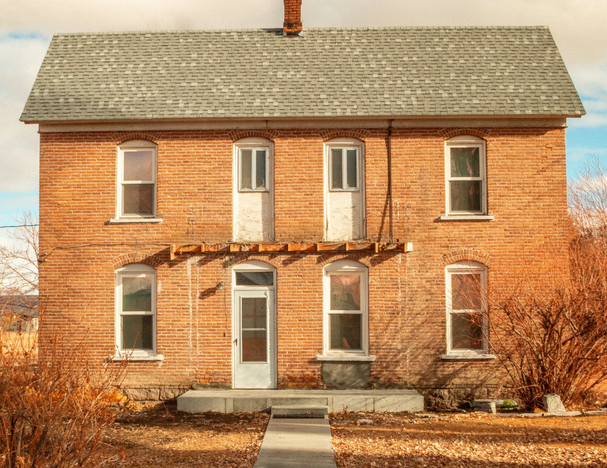 A two-story brick house with a pitched shingled roof, missing front porch, and boarded-up second-story doors. The house has six windows, three on each floor, with white trim. The front door is gray with a small window, and there are concrete steps le