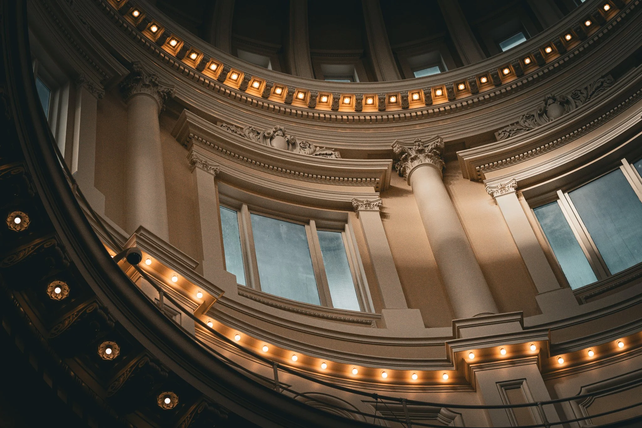 Interior view of a grand, circular building with multiple levels, featuring ornate architectural details, columns, large windows, and warm lighting along the railings.