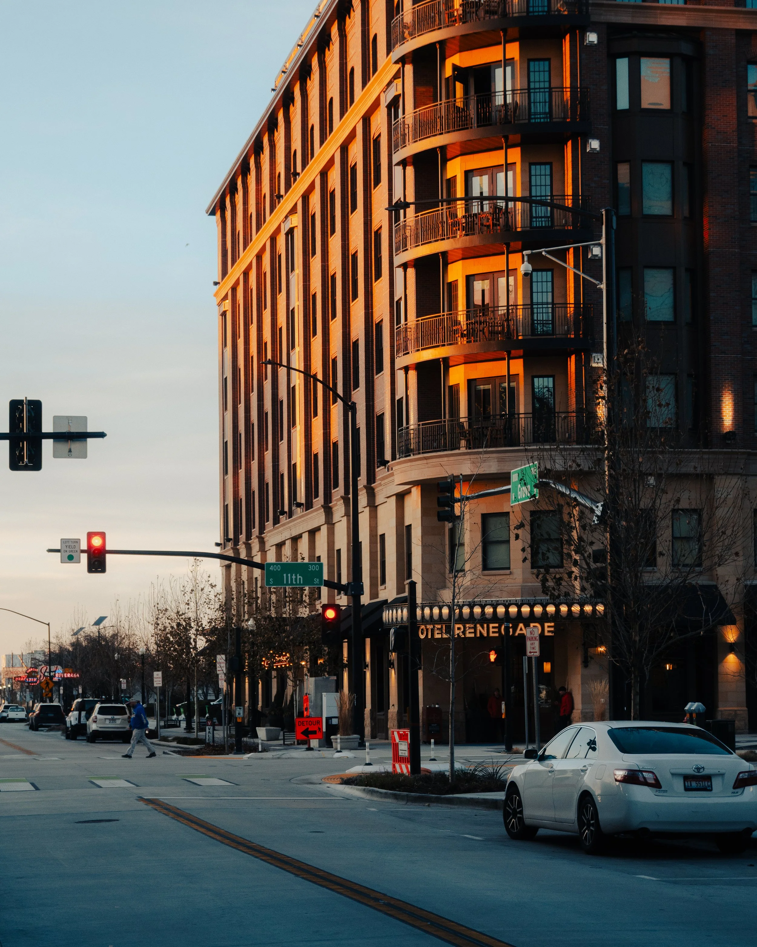 City street scene at sunset with a corner building illuminated by warm light, cars stopped at a red traffic light, pedestrians crossing, and a restaurant sign for 'Hotel Renegade' on the building.