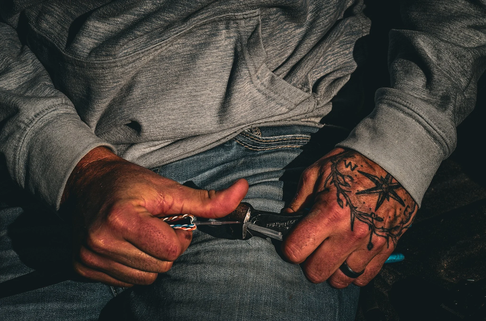 A person wearing a gray hoodie and blue jeans, with tattoos on their hands, is holding a small folding knife and cutting through a cable or wire.