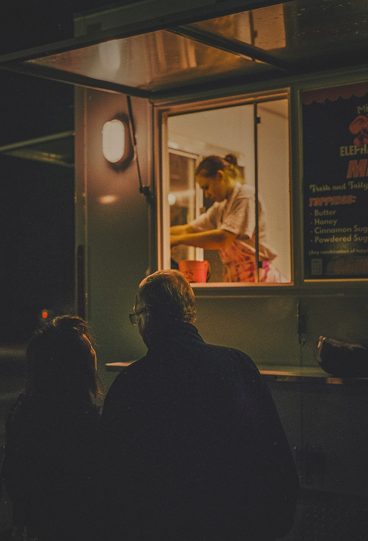 A food truck at night with a woman inside preparing food. Two people are sitting outside the food truck, one with glasses and the other with long hair, watching the woman through the window.