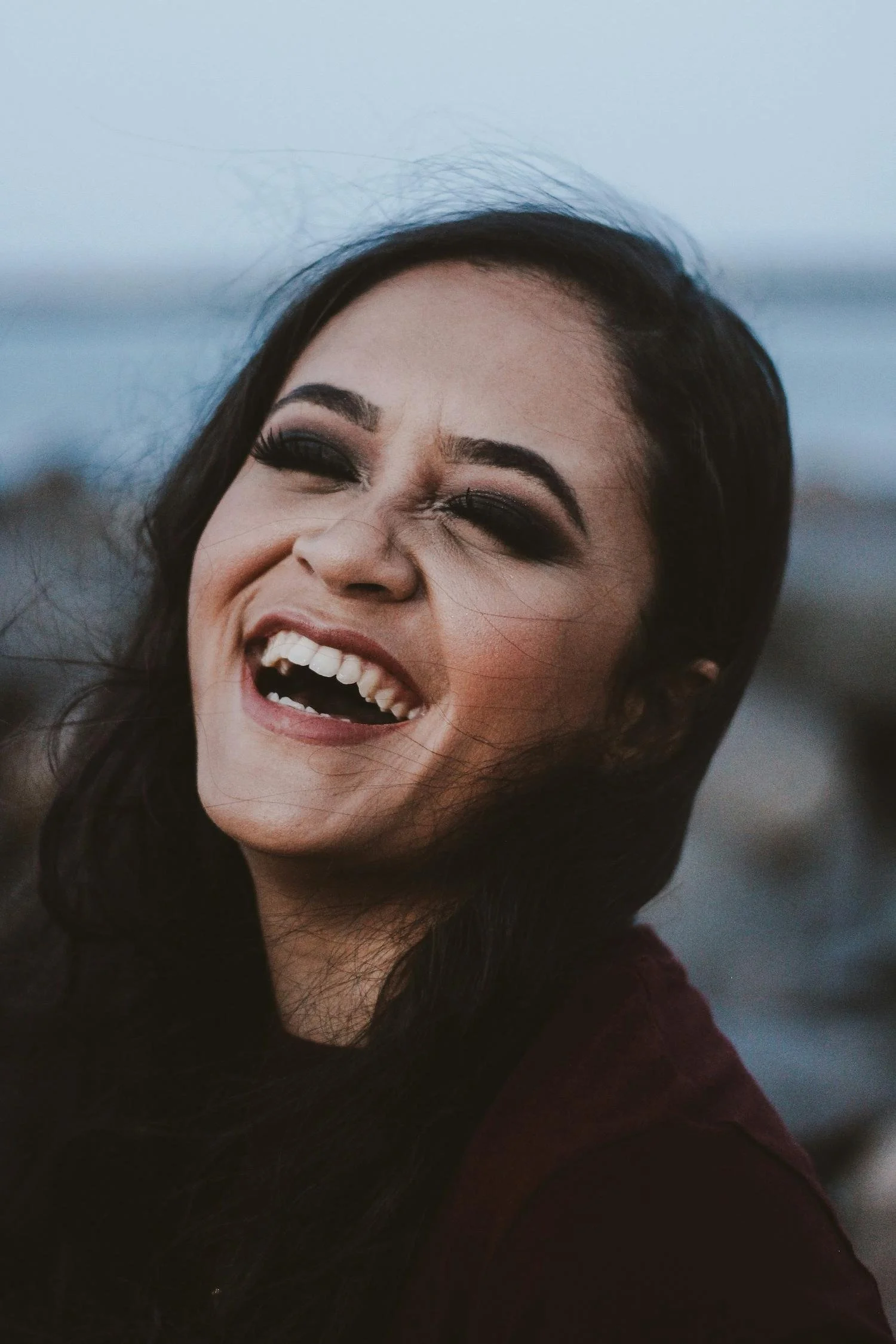 A woman with black hair and makeup smiling, showing her teeth, outdoors with a blurred background.
