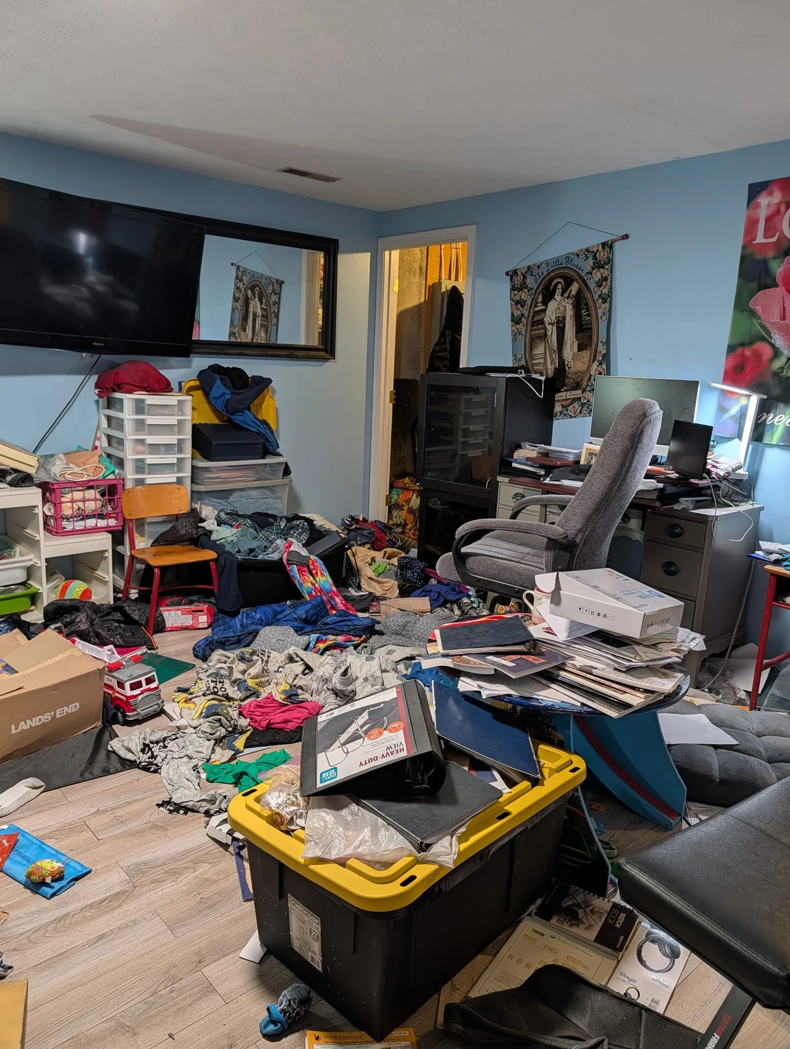 Room cluttered with clothes, boxes, and miscellaneous items, including a black trash bin with a yellow lid, a chair, a computer monitor, and various scattered objects on the floor.