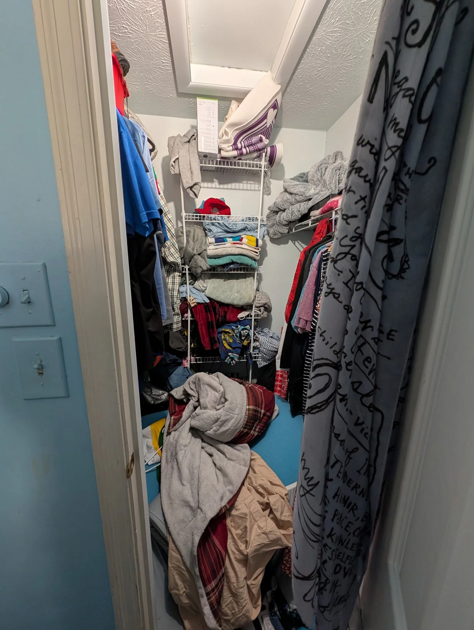 A cluttered walk-in closet with clothes and towels on wire shelves, a pile of blankets and clothes on the floor, and a wall-mounted mirror in the back wall.