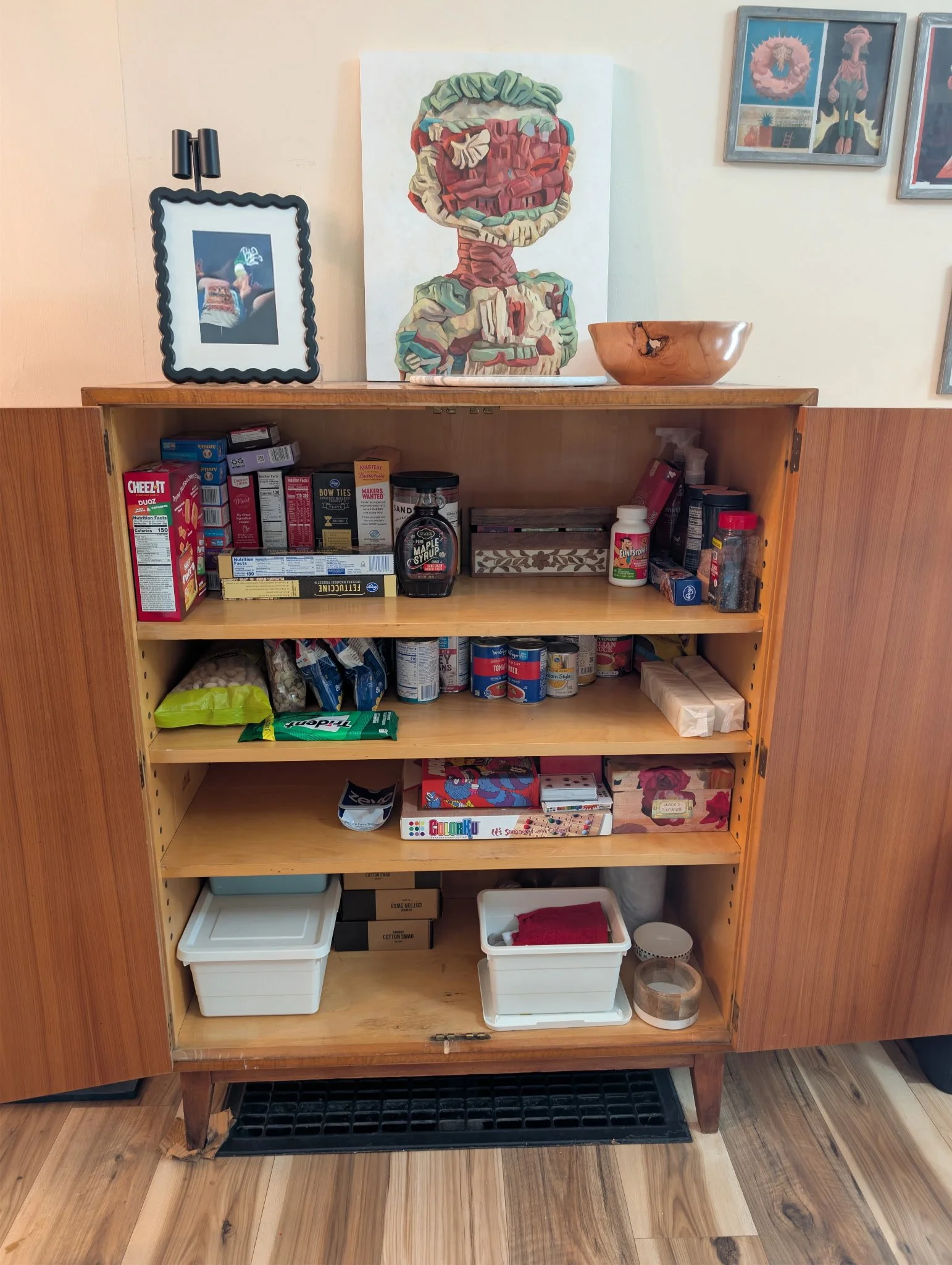 Organized wooden kitchen cabinet filled with various food items and kitchen supplies, with framed artwork on the wall above.