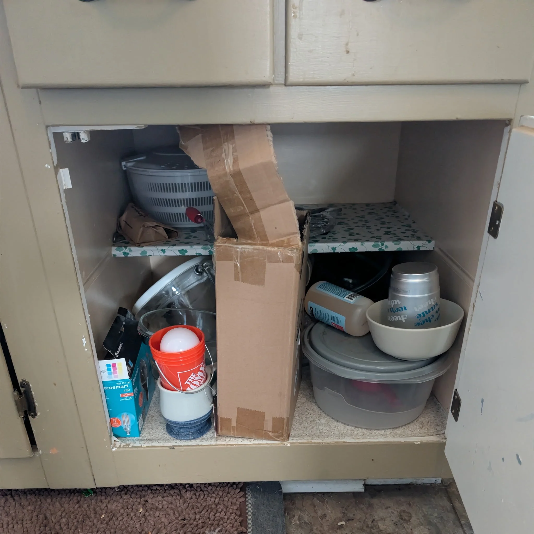 Open kitchen cabinet with various items, including a plastic food containers, a cardboard box, a paper bag, cleaning supplies, and dishes inside the lower shelf.