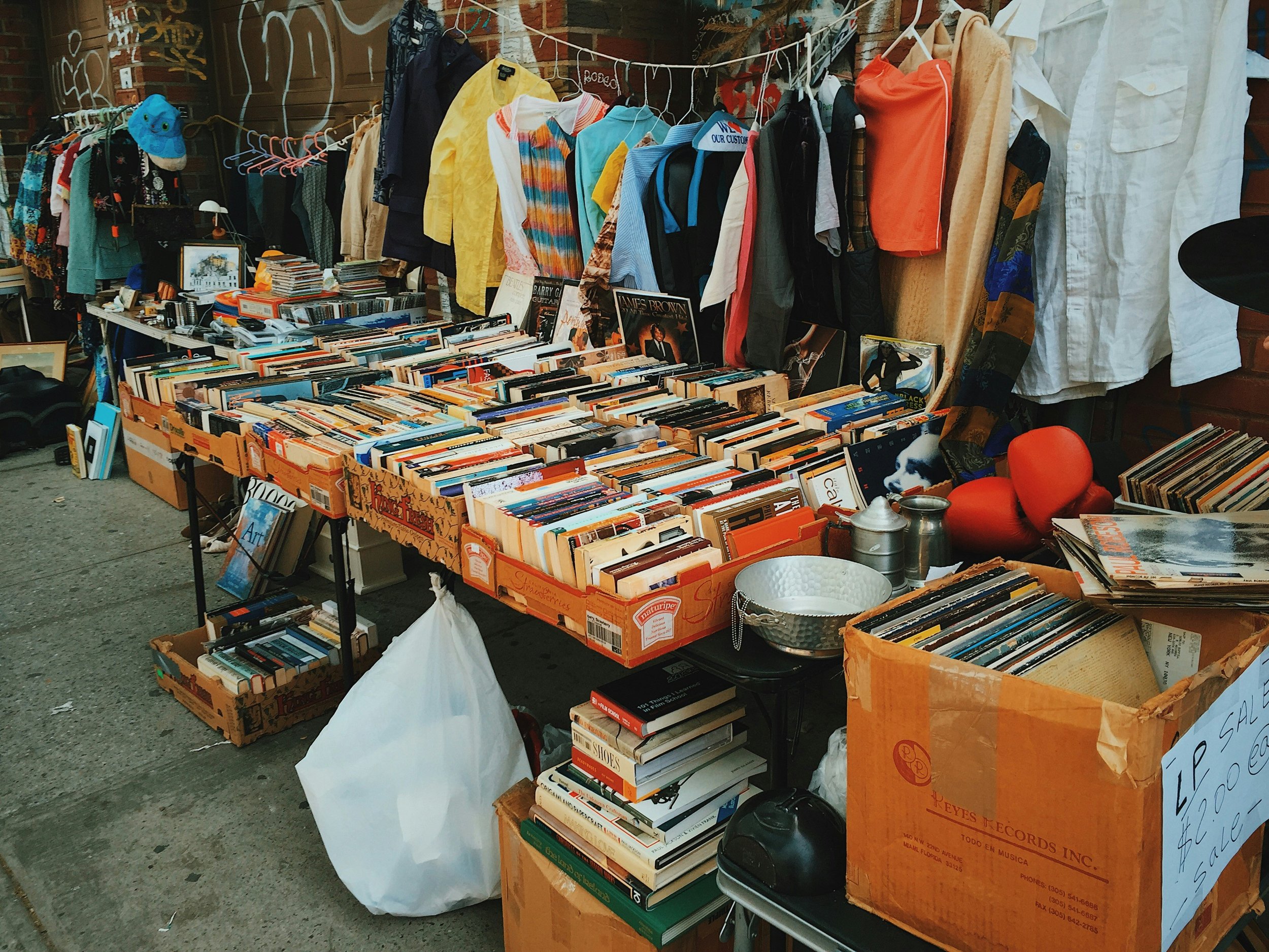 Various items at an estate sale or flea market. The center table holds wooden boxes filled with books. A box of vinyl records sits beside the table. Clothes hang behind the table from a clothing rack. Some items are stacked below the table.