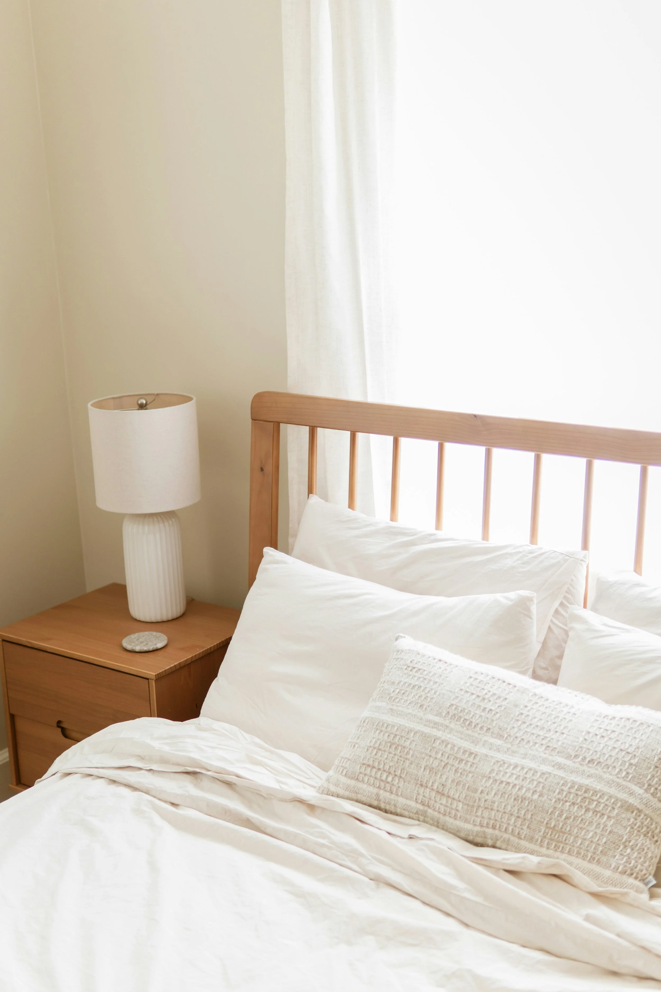 A bedroom with a bed made up with white bedding, pillows, a wooden headboard, and a nightstand with a lamp and a small stone. The room has white curtains and beige walls.