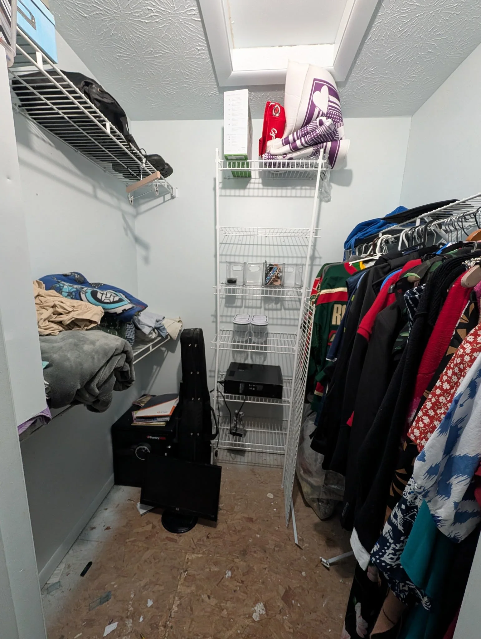 Small closet with clothing hanging on a rack on the right, a wire shelf on the left with folded clothes, and a small black safe on the floor.