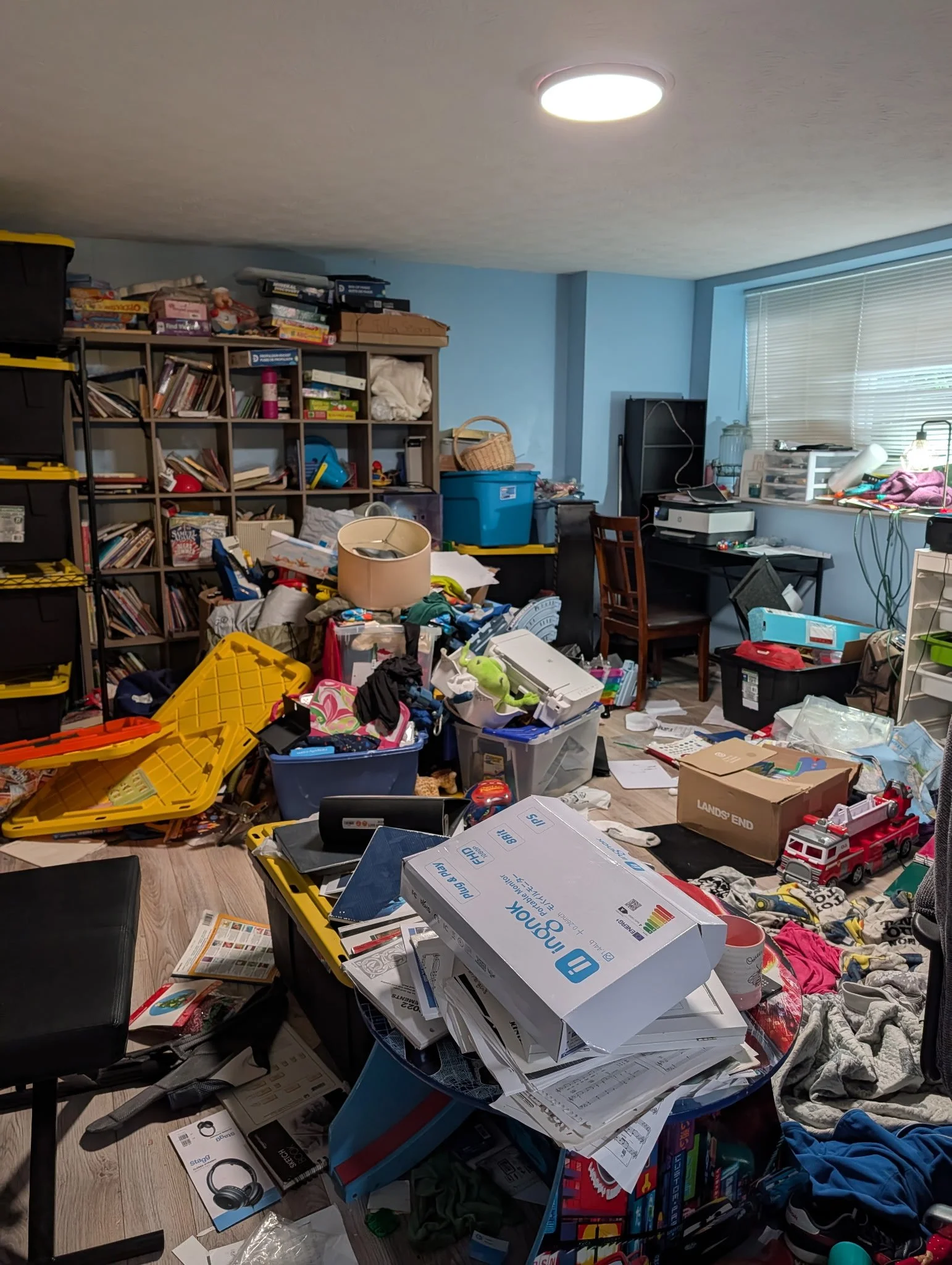 A cluttered room with a bookshelf filled with books and miscellaneous items, a table with disorganized papers, boxes, and supplies, a window with blinds partially open, and various toys and household items scattered across the space.
