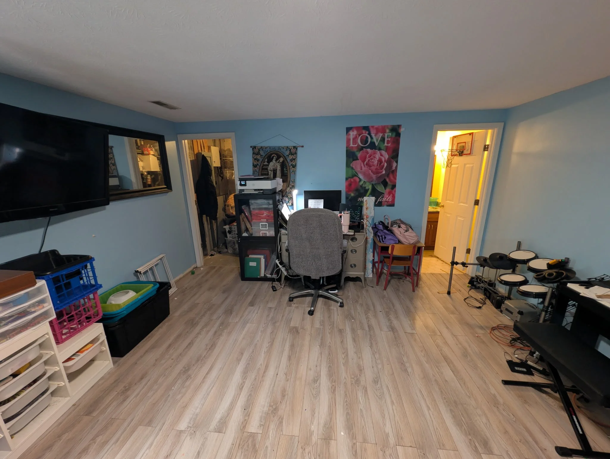 An organized room with wood flooring, a monitor, a treadmill, various furniture, artwork on the wall, and an open door showing a yellow-lit room.