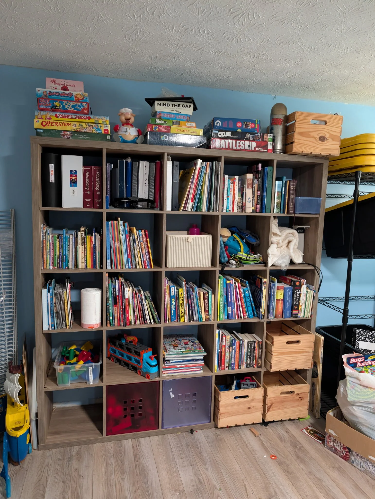A wooden bookshelf filled with books, toys, games, storage bins, and miscellaneous items in a room with wooden flooring and a blue wall.