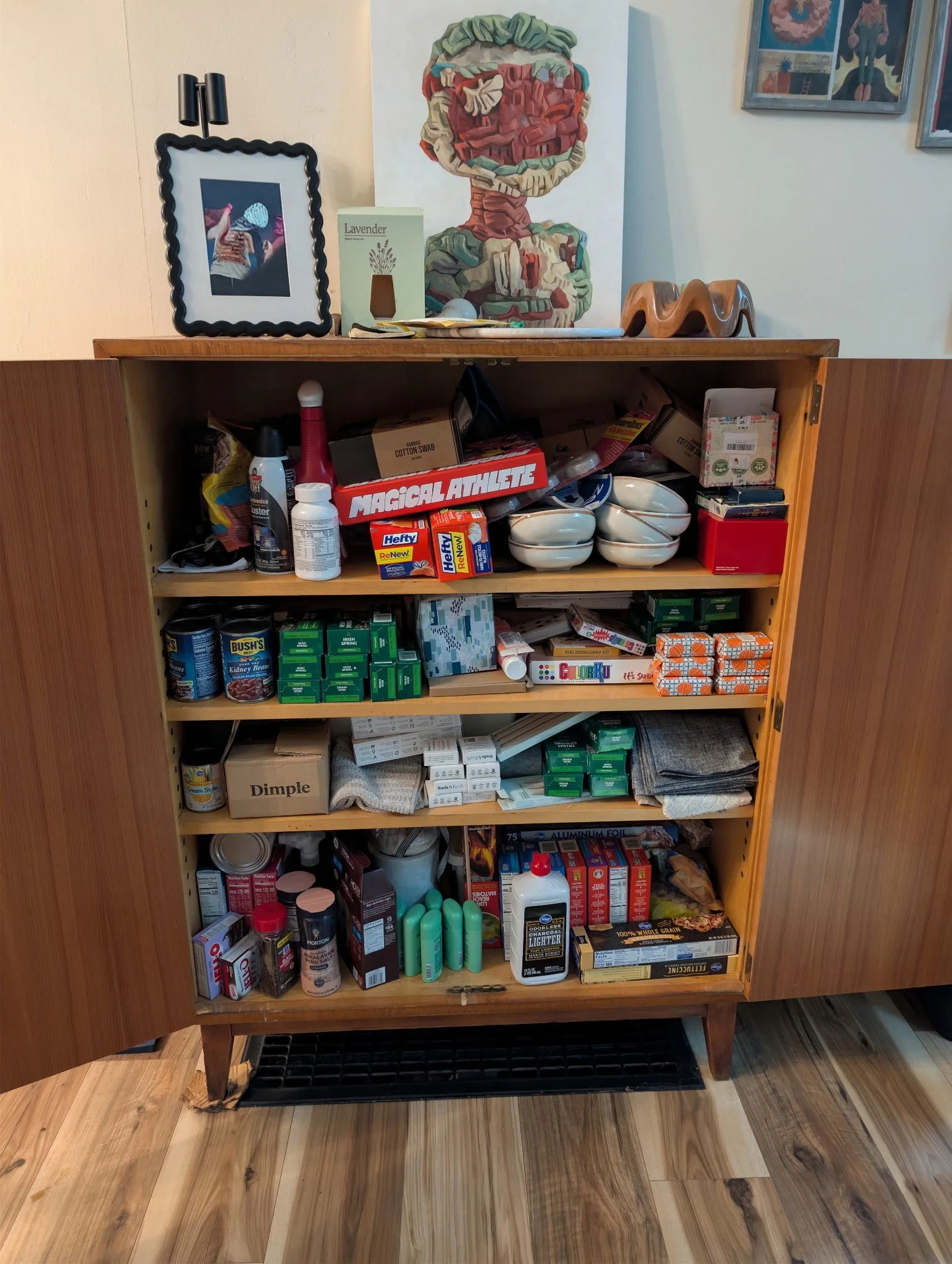 A cluttered wooden cabinet with open doors filled with various household supplies, including cans, boxes, bottles, and cleaning products, with a hardwood floor in front and artwork on the wall above.