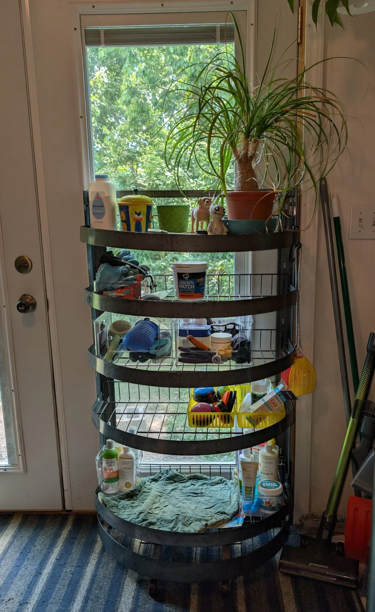 A vertical metal shelving unit with six tiers positioned in a room near a door with a window, holding various household items including skincare products, tools, a container, a cloth, and a potted plant, with a view of trees outside the window.