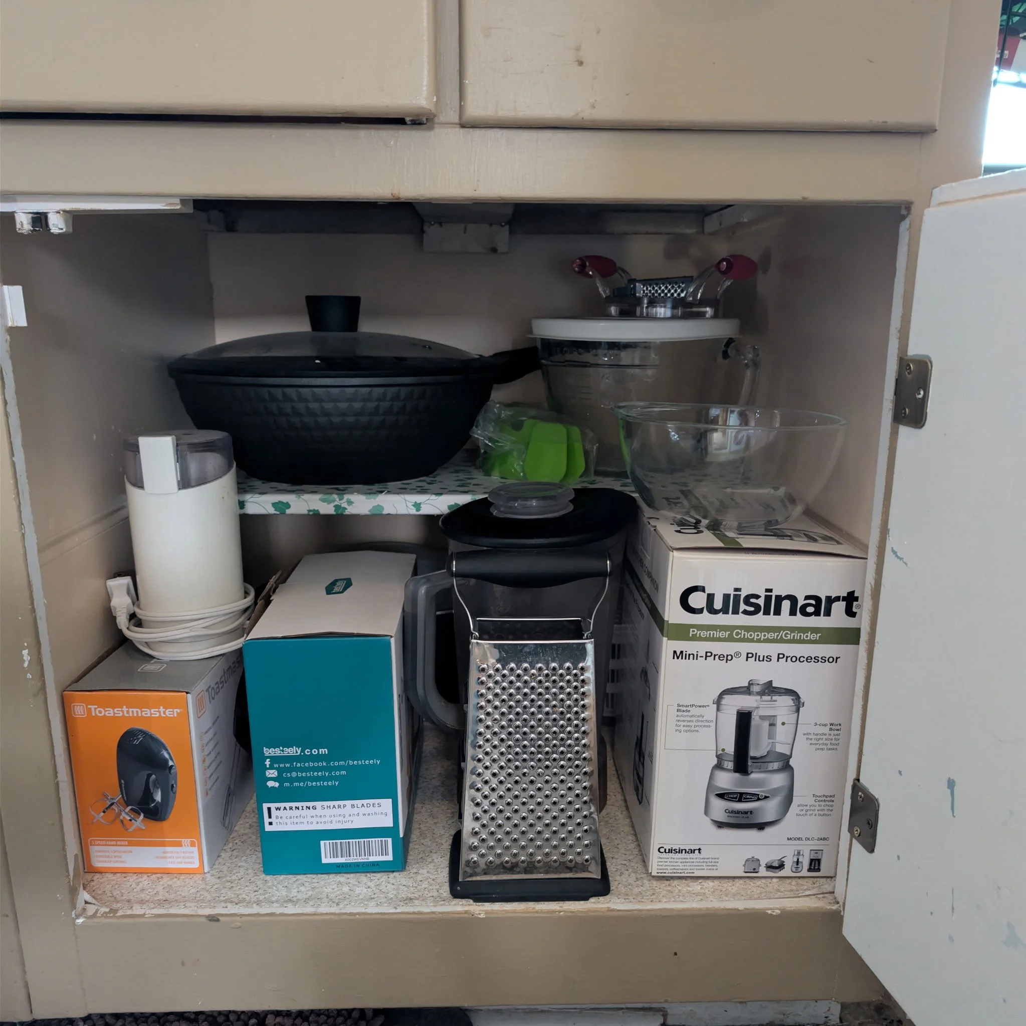 Underneath a kitchen cabinet, various small appliances and boxes are stored, including a Cuisinart mini-prep processor box, a Toastmaster appliance, and a Besteely food processor box. There is also a black bowl, a metallic grater, and a clear glass bowl among the stored items.