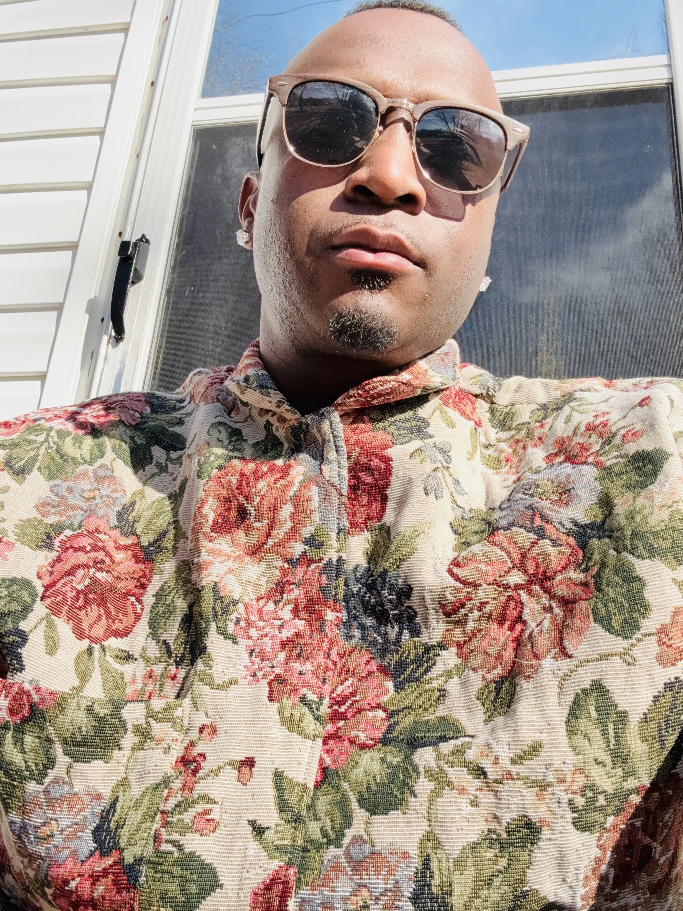 A person taking a selfie outside, wearing sunglasses and a floral-patterned shirt, with a house and blue sky in the background.