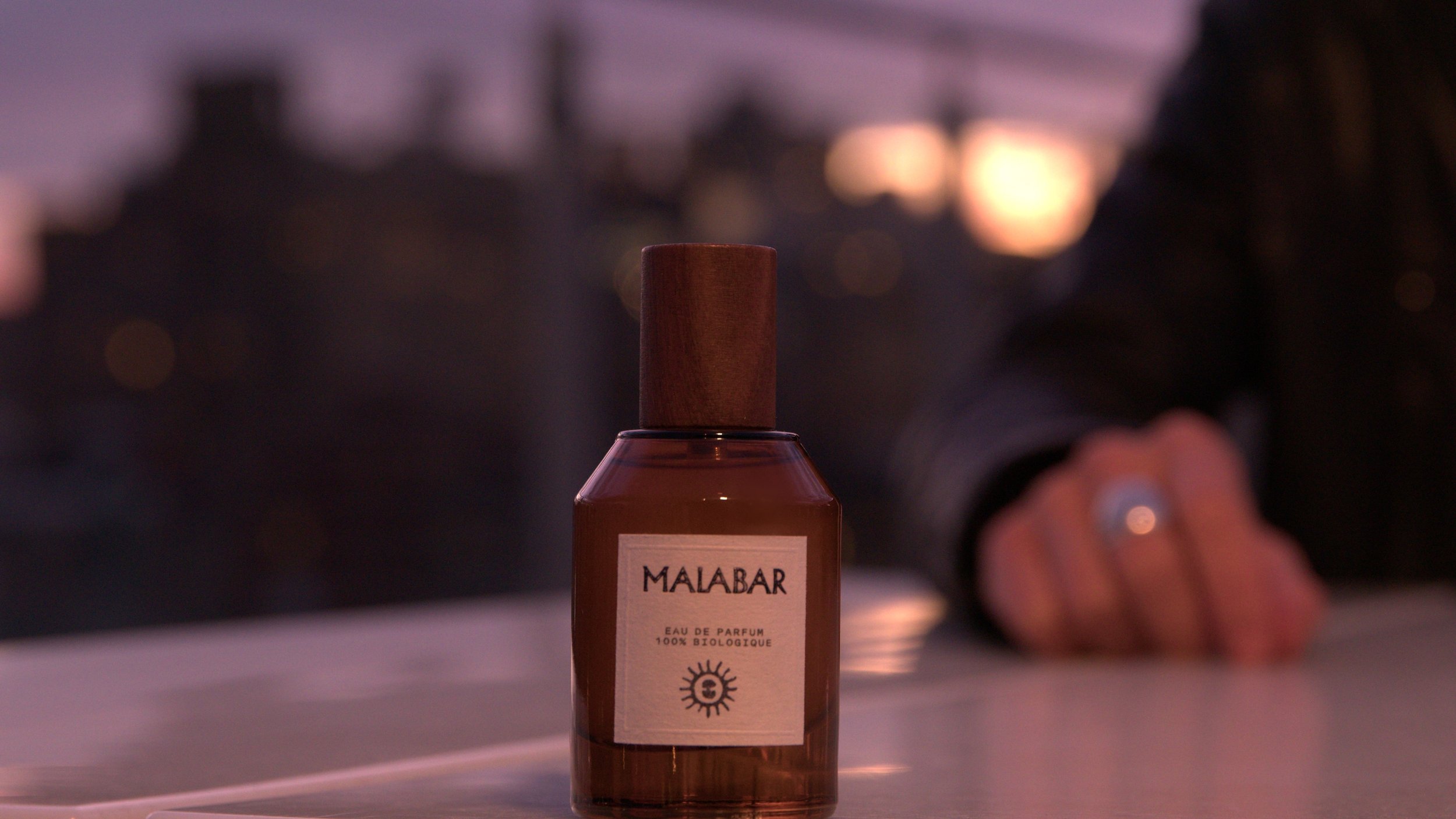 A bottle of Malabar Eau de Parfum with a wooden cap on a reflective surface, with blurred city lights and sunset in the background.