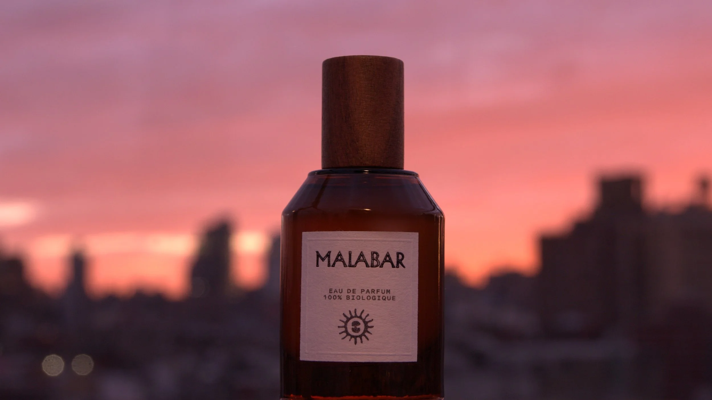 A bottle of Malabar perfume with a wooden cap, set against a blurred city skyline at sunset with pink and purple clouds.