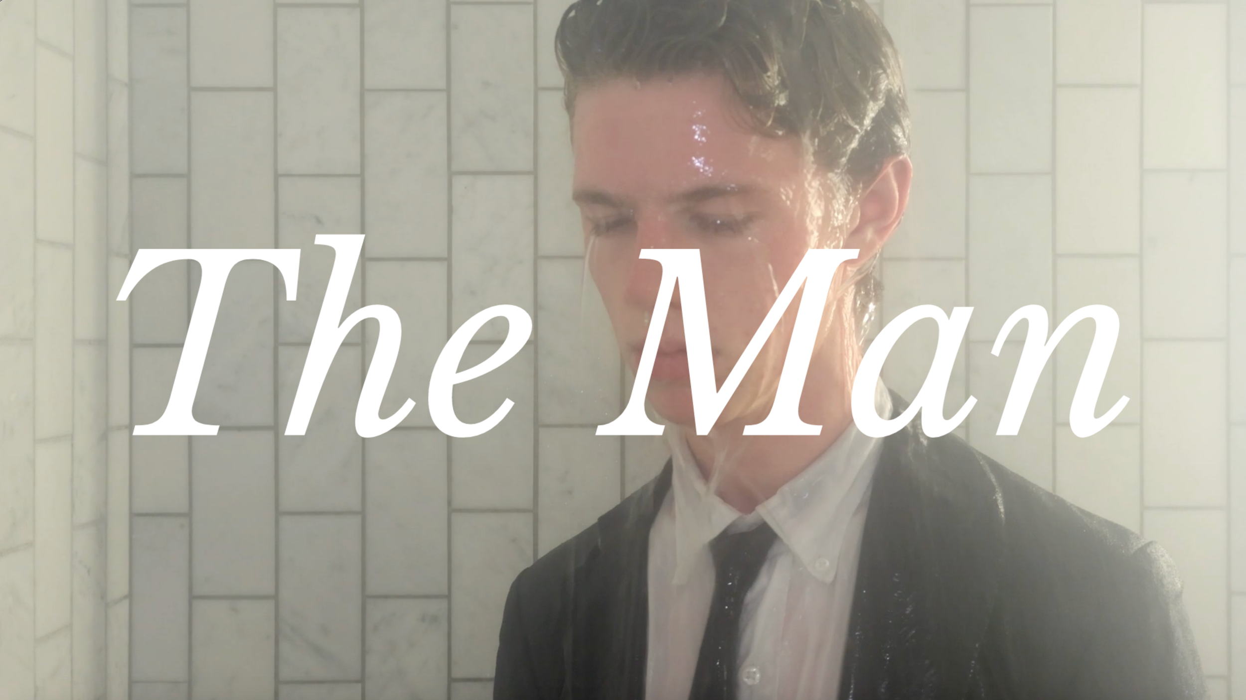 Person with wet hair, wearing a white dress shirt and black blazer, standing in a tiled shower. The words "The Man" are overlayed on the image.