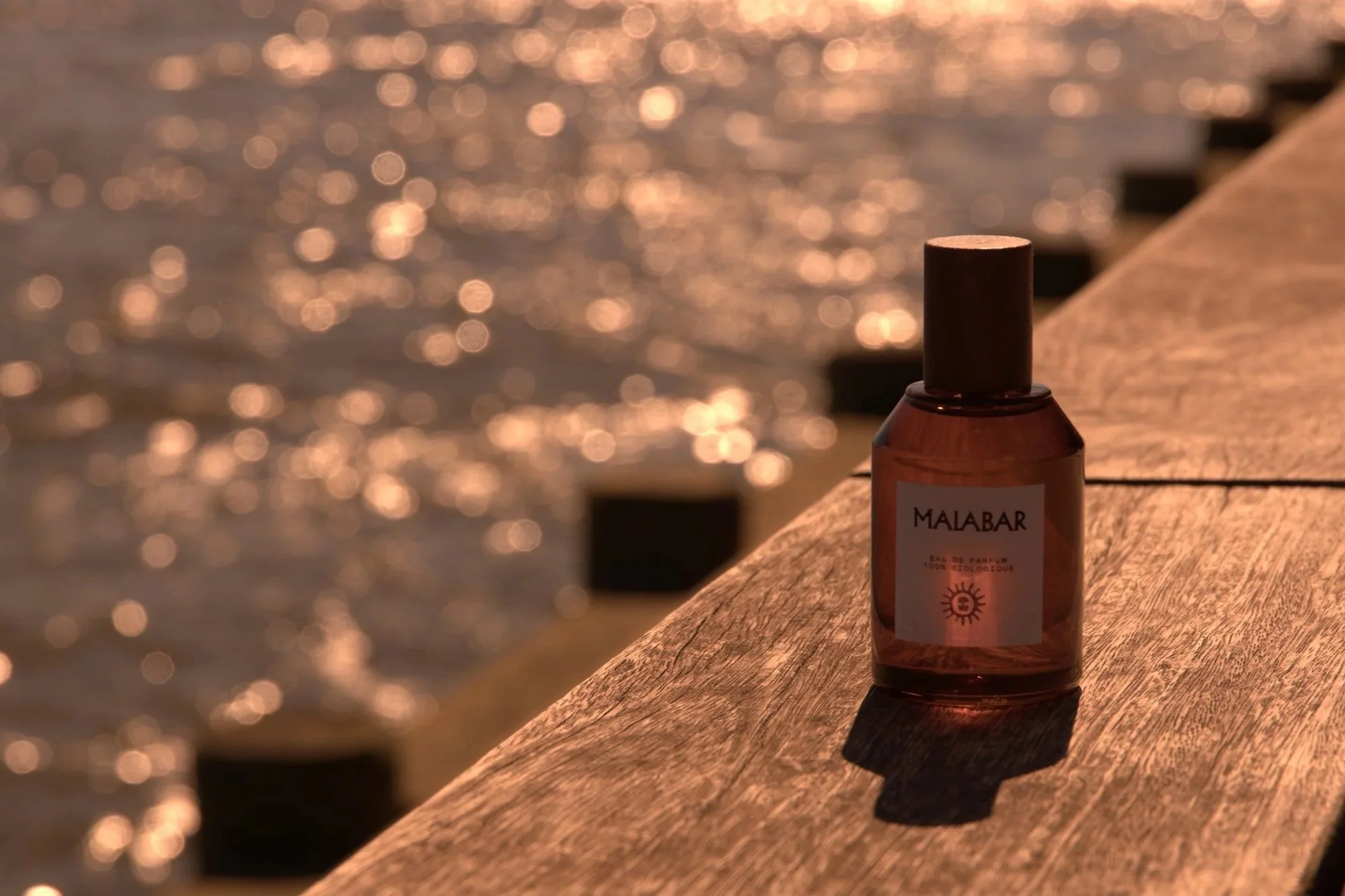 A small bottle of perfume labeled 'MALABAR' placed on a wooden surface near the water with sunlight reflecting off the waves in the background.
