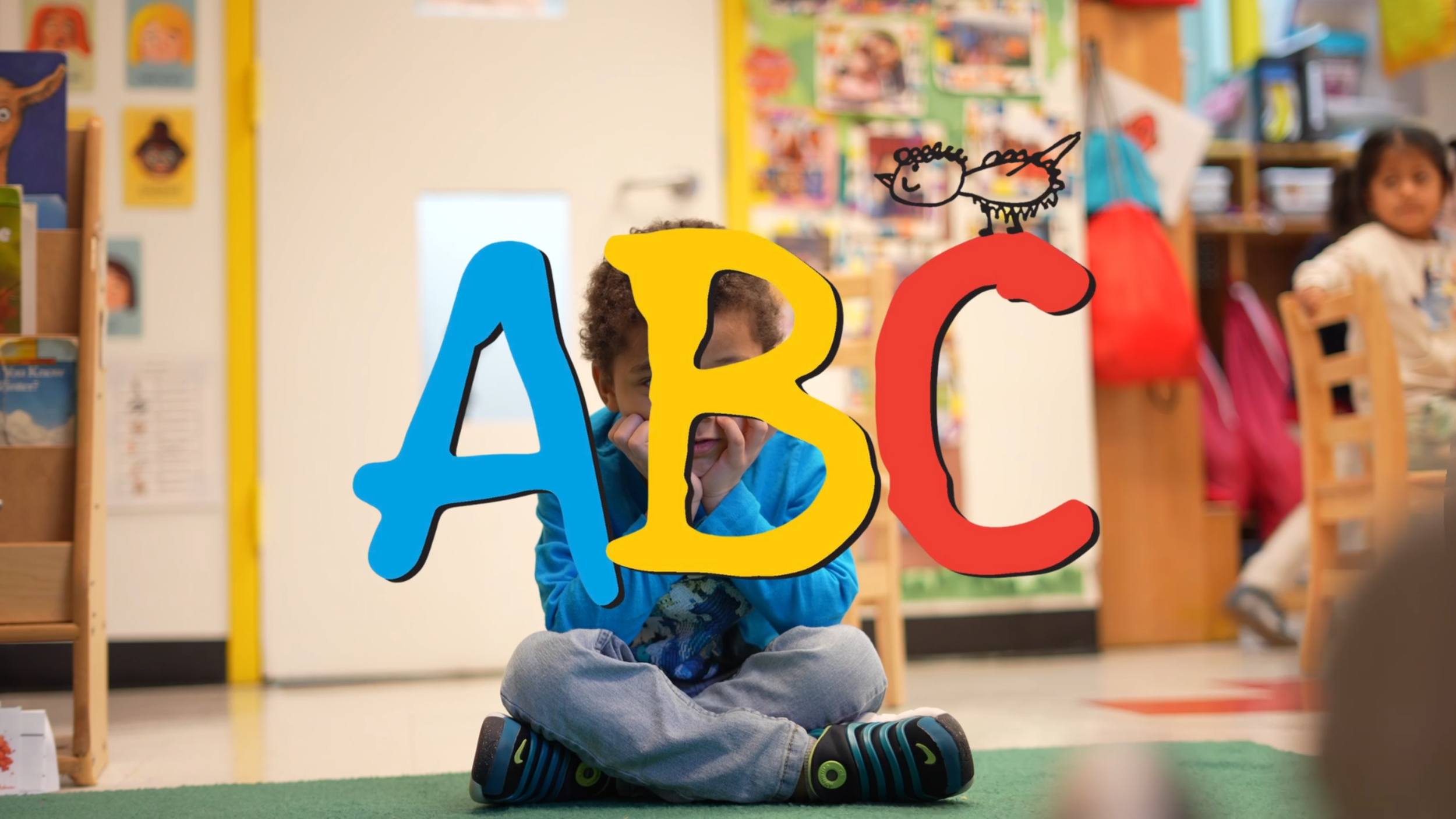 A young boy with curly hair sitting cross-legged on the floor in a classroom, looking through a colorful cutout of the letters A, B, and C with a small drawing of a unicorn on the letter C. Two other children are visible in the background, along with bookshelves and colorful classroom decor.