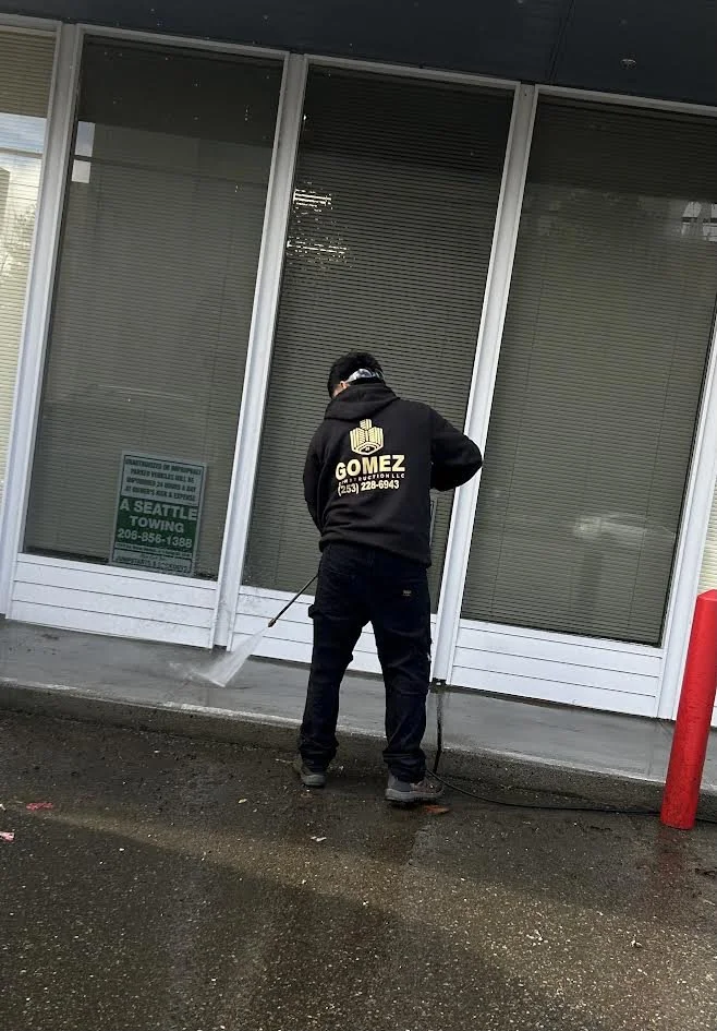 A person power washing the sidewalk outside a building with large glass windows and vertical blinds, wearing a black hoodie with a logo and contact information on the back.