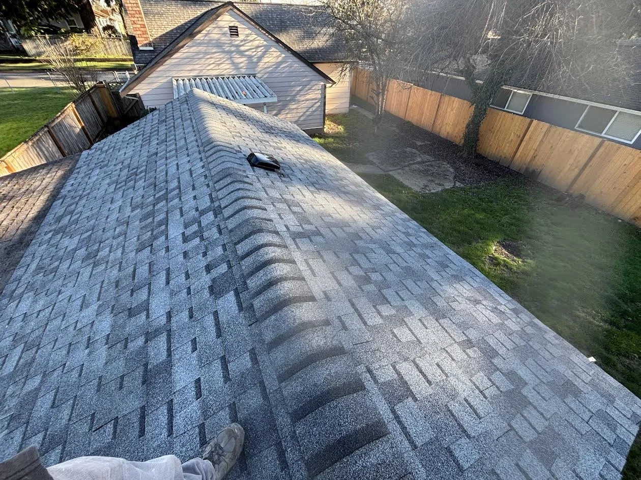 View from the roof of a house showing gray asphalt shingles, a vent, and a person's foot in the bottom corner. The yard is fenced with wooden panels, with a smaller shed and some trees visible.