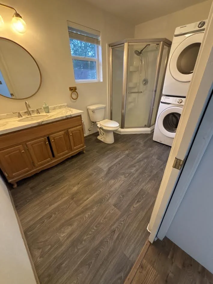 Bathroom with wooden vanity, marble countertop, round mirror, toilet, corner shower with glass doors, stacked washer and dryer, and window.