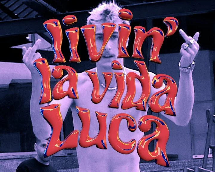 A shirtless man with platinum blonde hair holding up both middle fingers in front of a sign that reads 'living la vida LUCa' with colorful, shiny, 3D text.