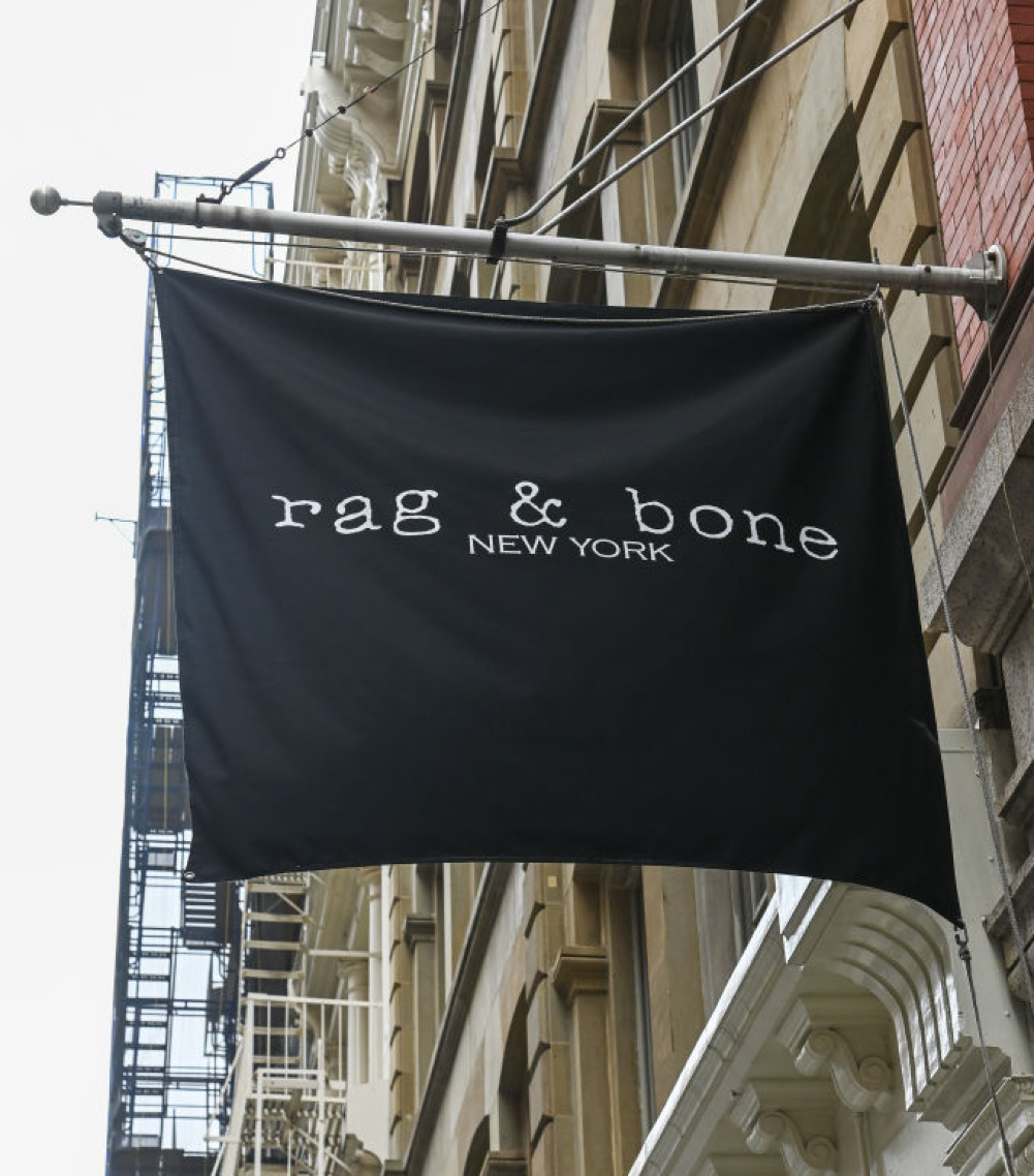 Black flag with white text reading "rag & bone NEW YORK" hanging from a building on a city street.