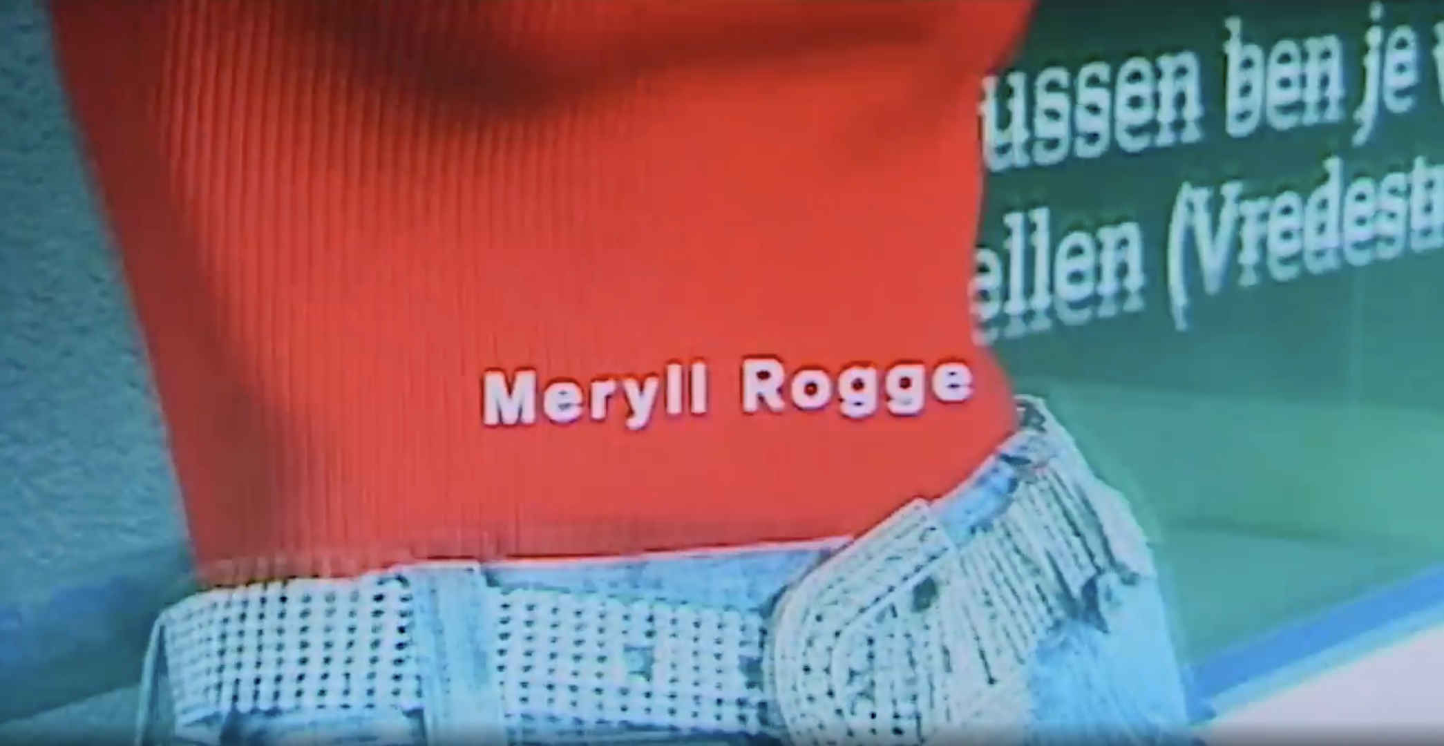 Close-up of a person wearing a checkered shirt, with a red bag or item bearing the name 'Meryll Rogge' in white text. Behind, part of a green surface with white readable text, and a textured blue background.