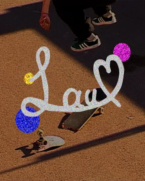 Colorful glittery decoration with the word 'Love' and two circles attached, placed on a skateboard on a beige carpeted floor. Part of a person wearing black pants and sneakers is visible.