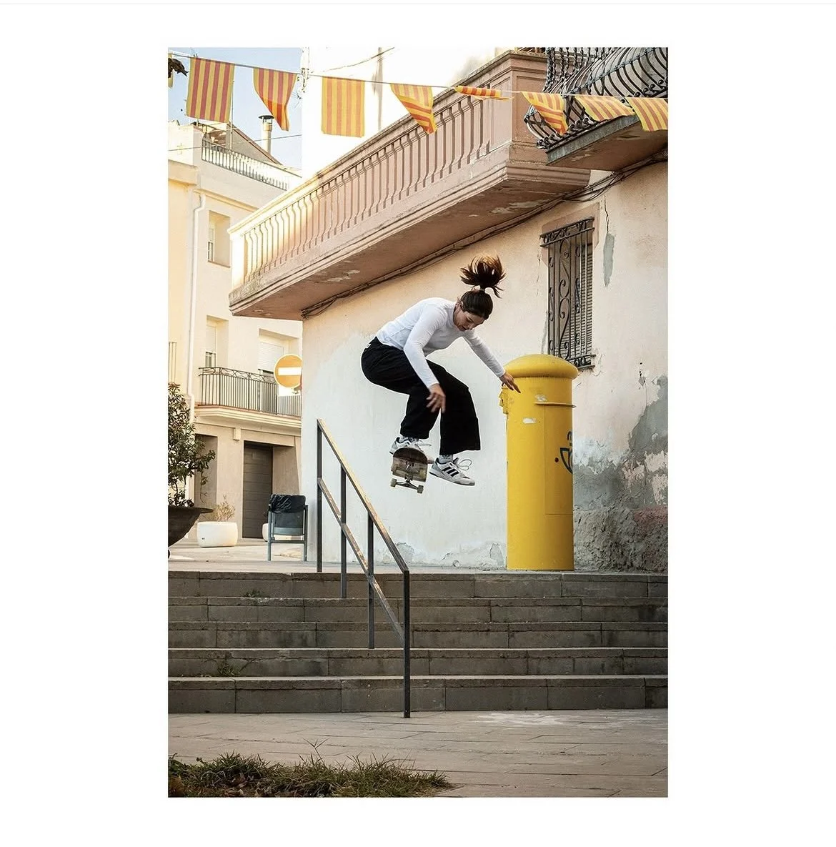 A woman skateboarding down a set of stairs and jumping over a yellow mailbox on an urban street.