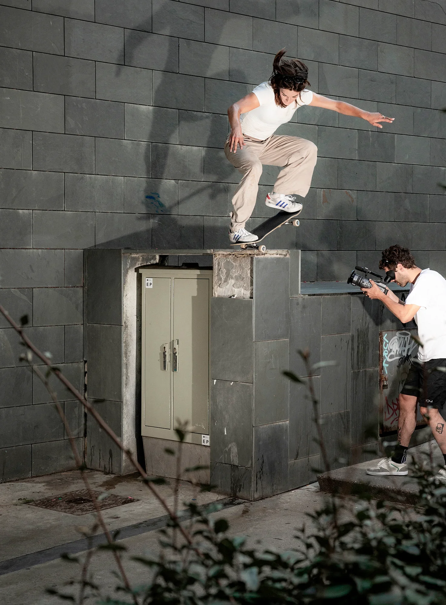 A woman skateboarding on a ledge in front of a gray brick wall. A person in white shorts and a white shirt is taking photos nearby. There are small plants in the foreground and some graffiti on the wall.