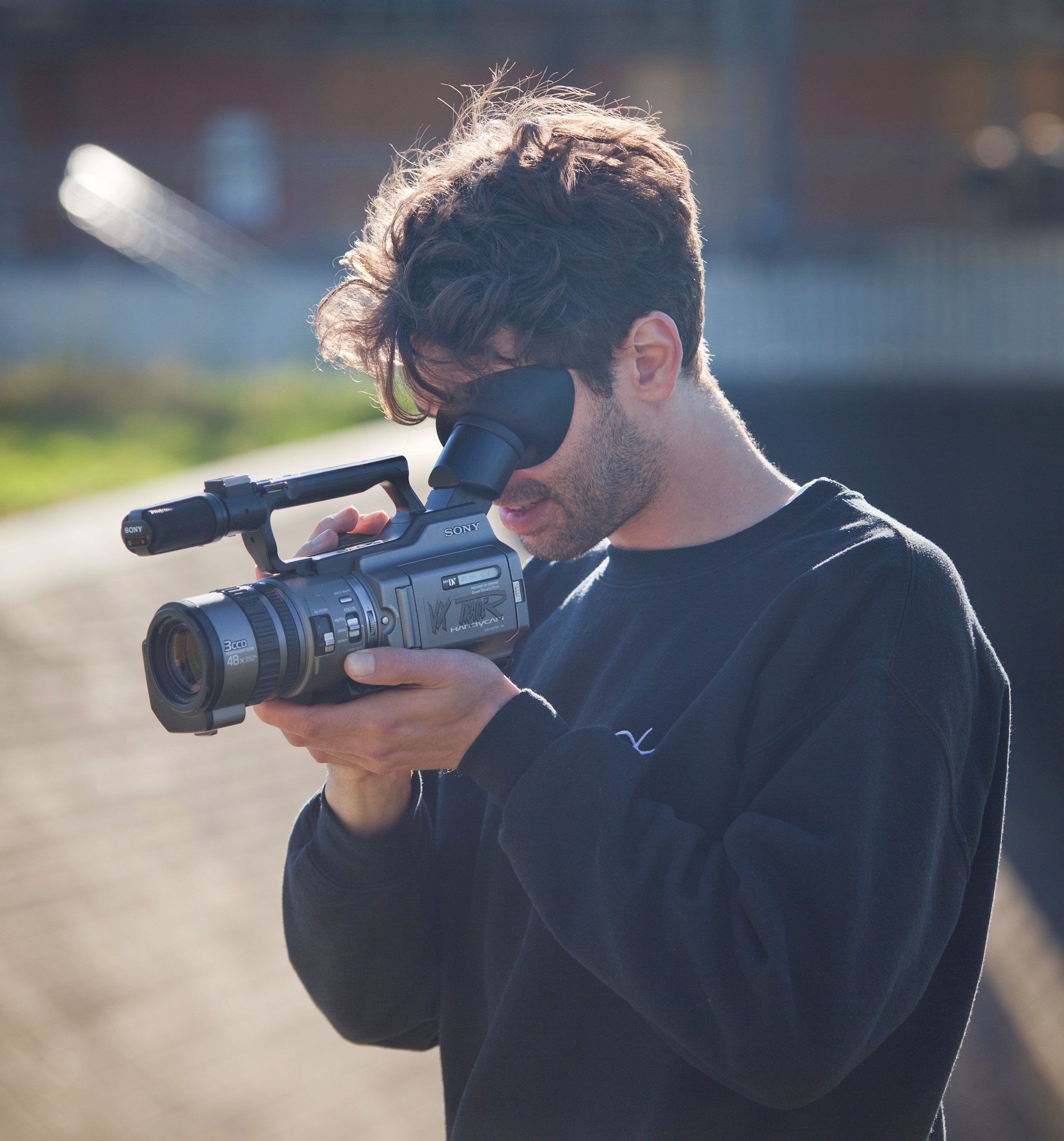A young man with curly hair and a beard looks through a video camera with a viewfinder, outside during daytime.