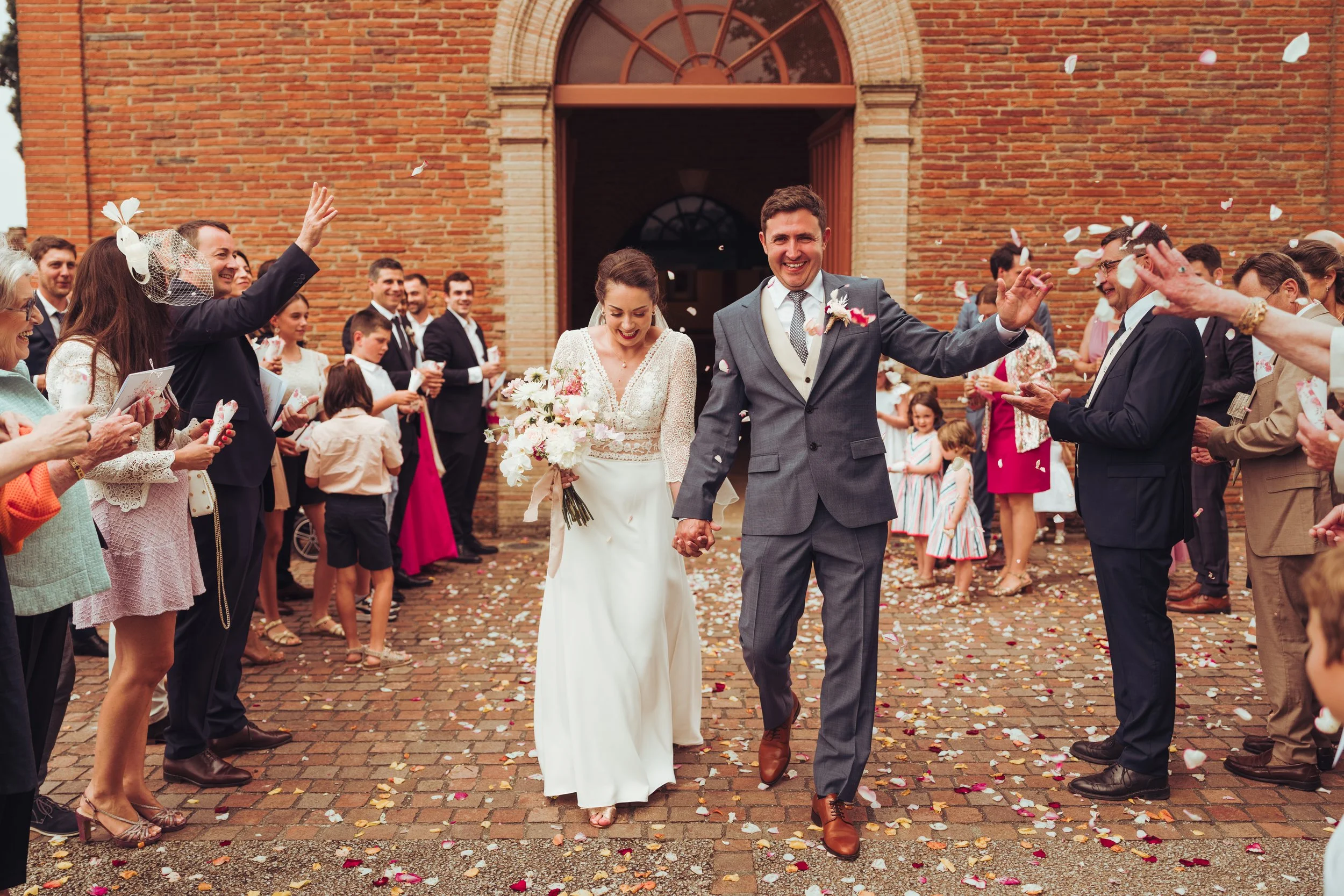 Un couple marié, lui en costume gris et elle en robe blanche, marche main dans la main sous une arche de fleurs, entouré de leurs invités lançant des pétales de rose et des confettis lors de la sortie de cérémonie religieuse proche de Toulouse.