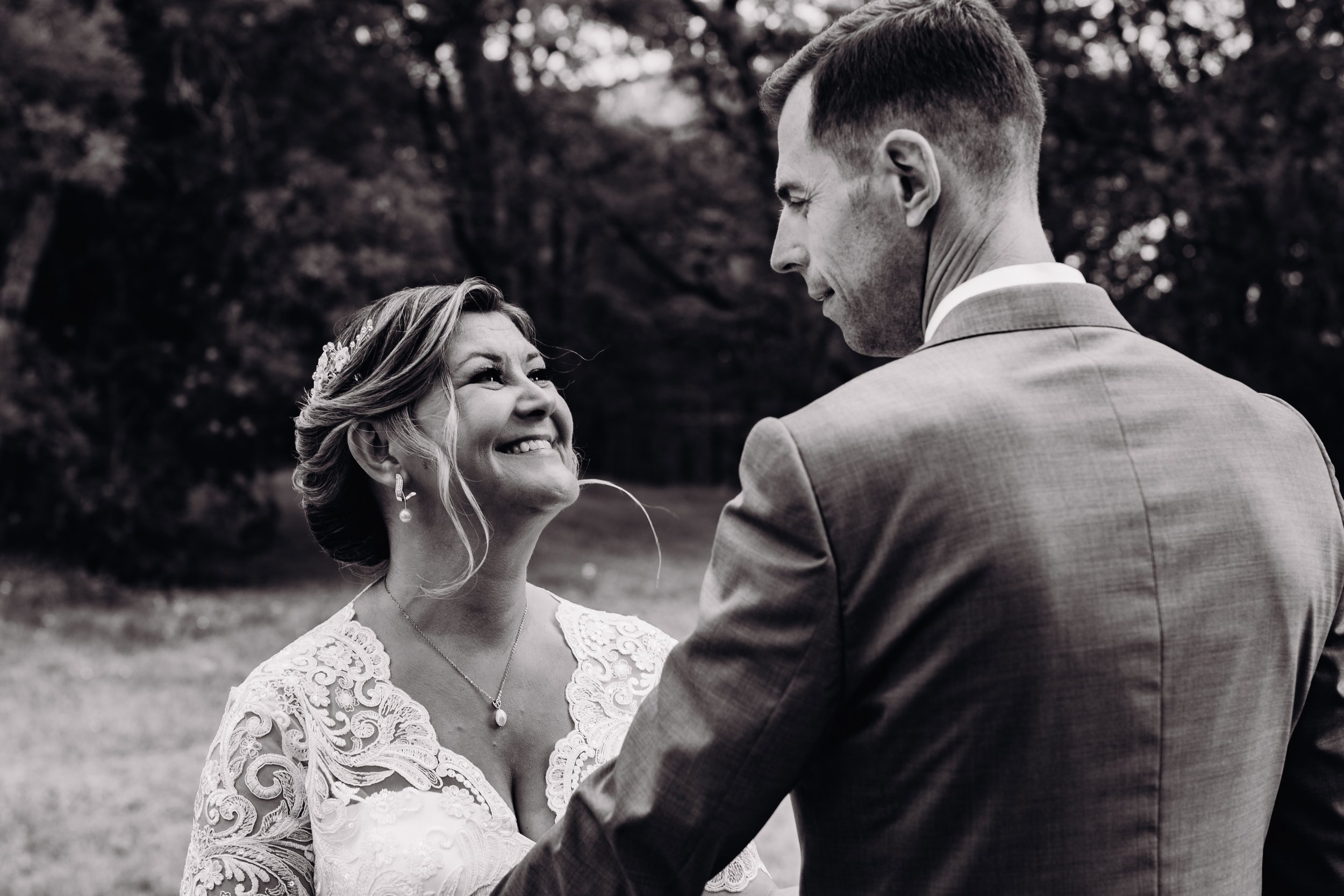 Une femme en robe de mariée sourit émotionnellement en regardant un homme en costume lors d'un mariage en plein air, en noir et blanc, proche de toulouse