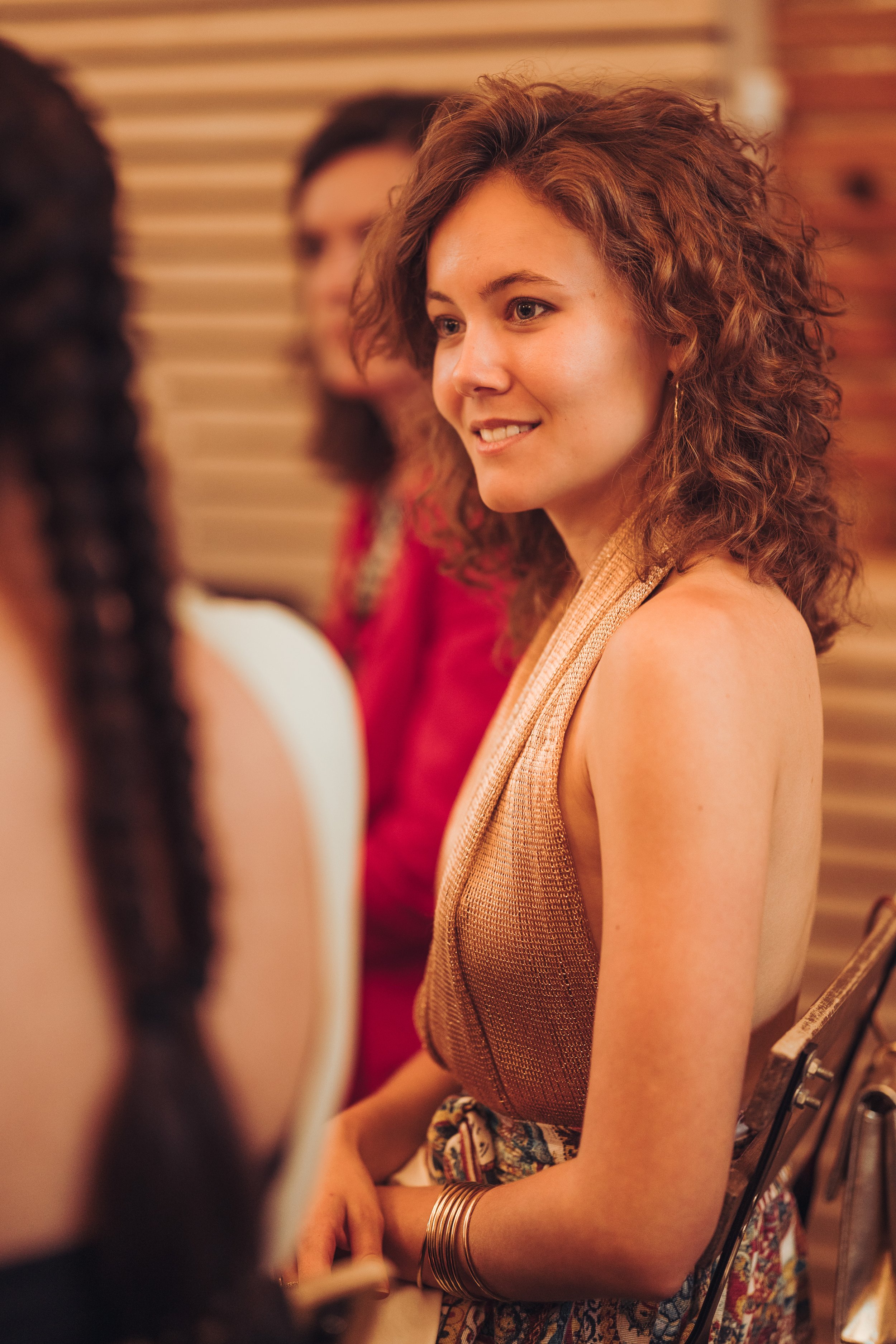 Une jeune femme avec des cheveux bouclés bruns, souriant, assise lors d'un évènement social, entourée d'autres personnes au domaine de Gailhaguet, proche de Toulouse, en Occitanie