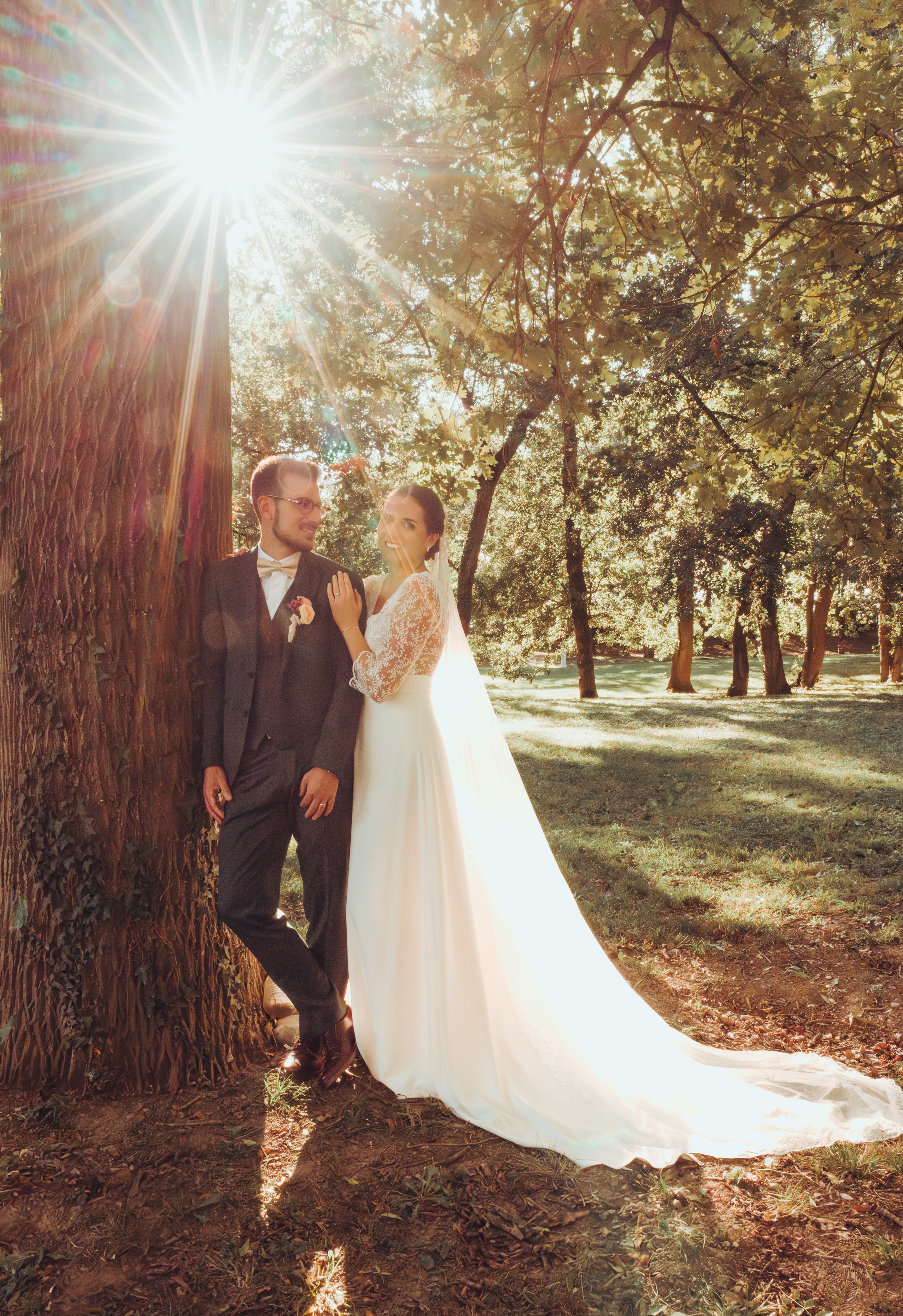 Un couple en robe de mariage debout près d'un arbre dans le parc du Moulin de Rudelle, sous un ciel ensoleillé avec des rayons de soleil lumineux.