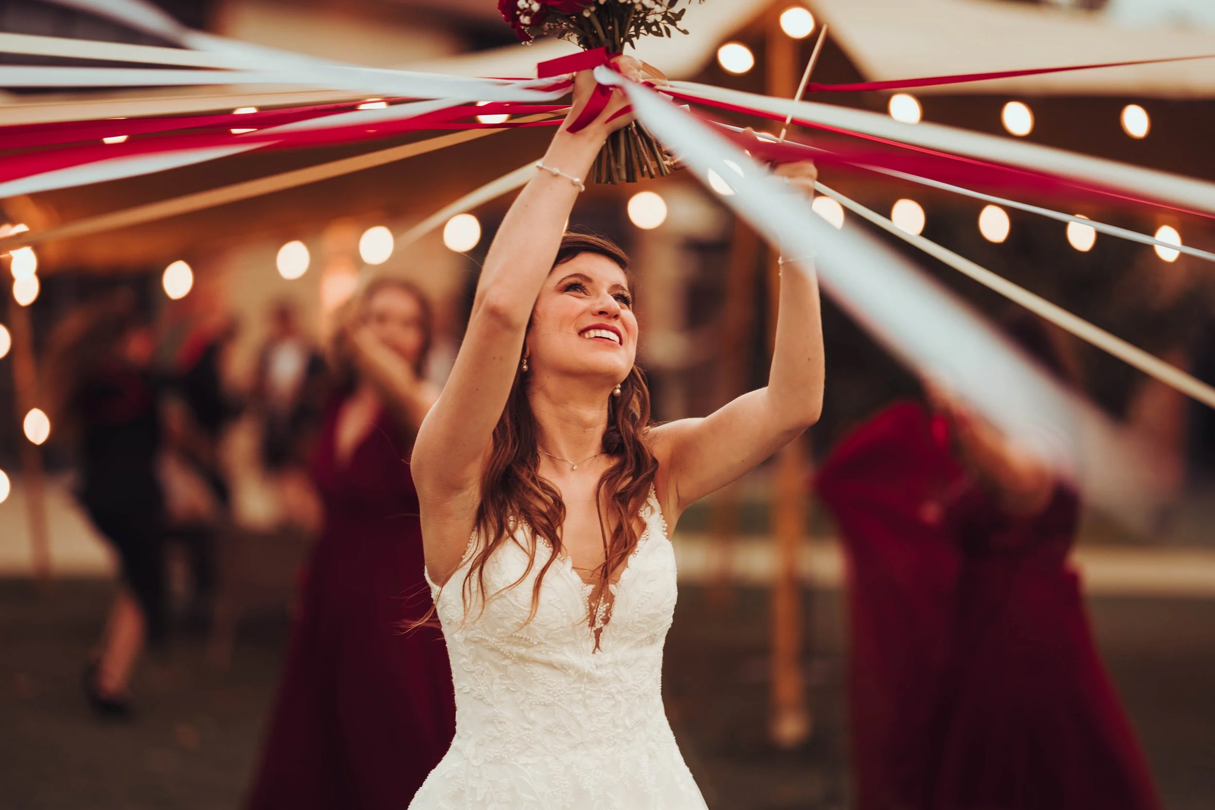 Une mariée réalise le jeu des Rubans lors d'une réception de mariage au Mas Tolosa proche de Toulouse, en Occitanie, entourée d'invités, sous des lumières décoratives, tenant un bouquet.