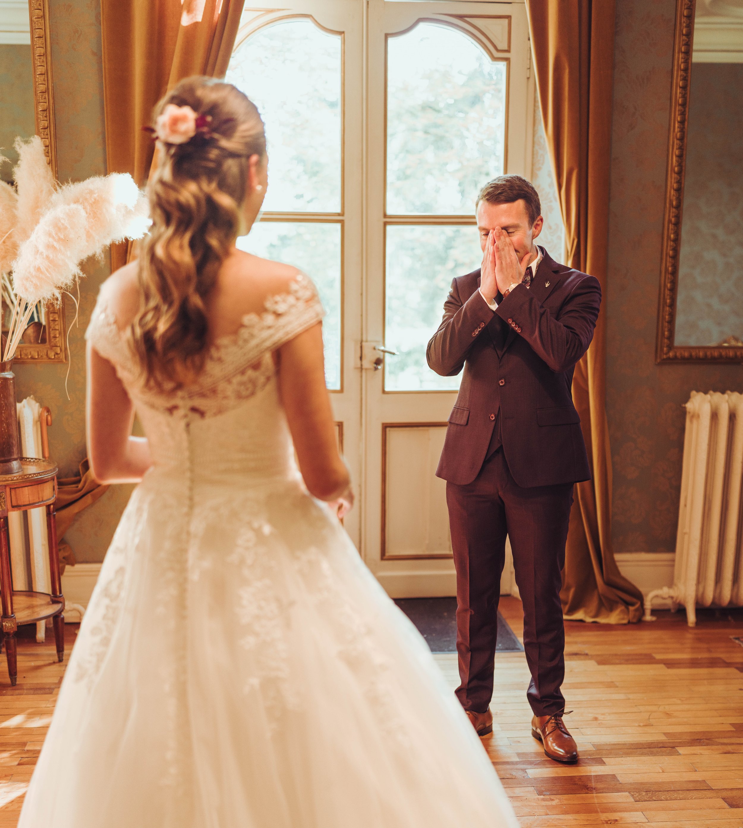 Un homme en costume regarde une femme en robe de mariage, avec les mains sur le visage, dans une pièce élégante avec des fenêtres et des rideaux dorés.