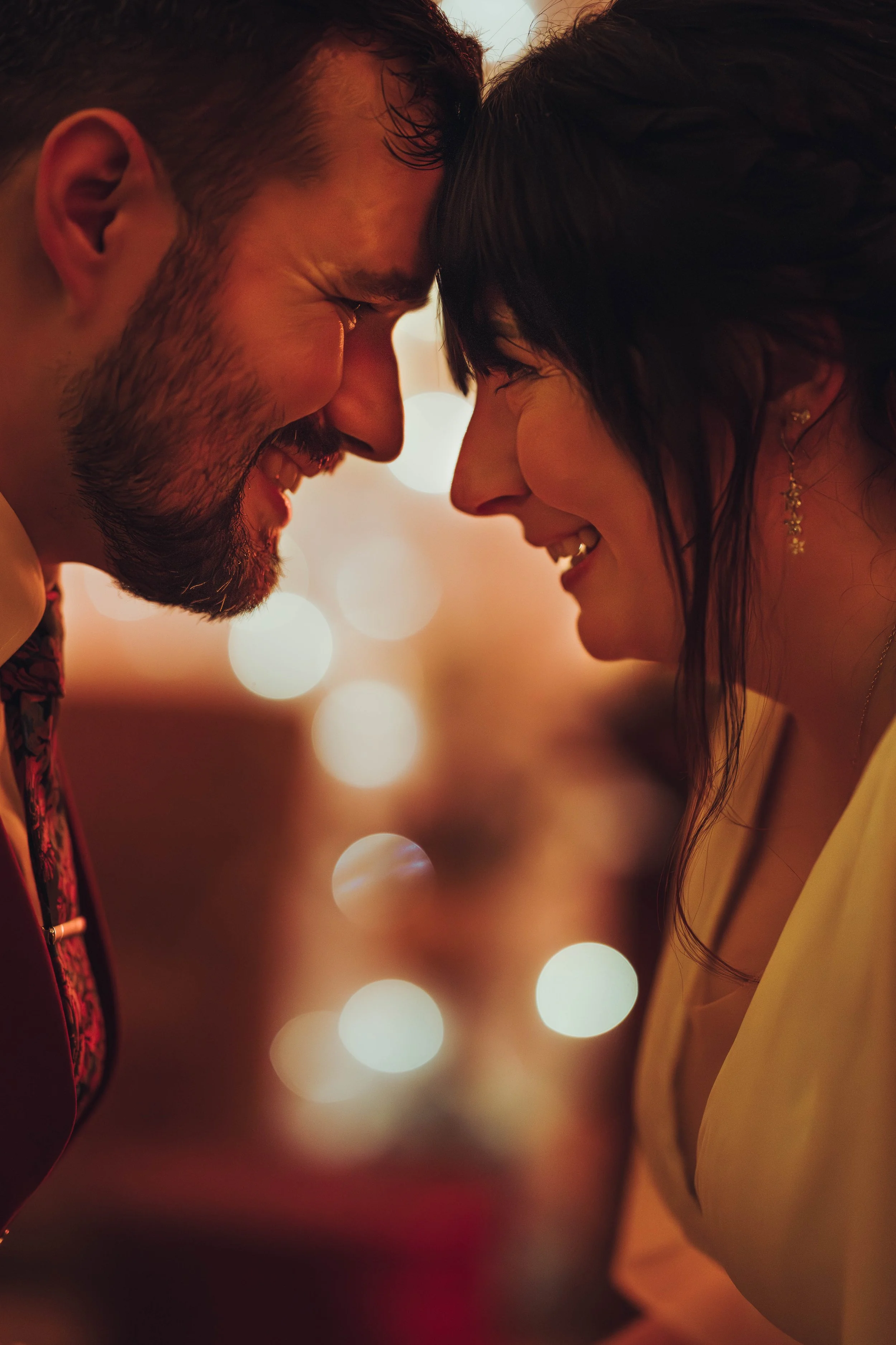 Un couple souriant, très proches, avec le front touchant, matière d'intimité et de bonheur, dans un décor chaleureux avec des lumières floues en arrière-plan au domaine de Gailhaguet proche de Toulouse. 