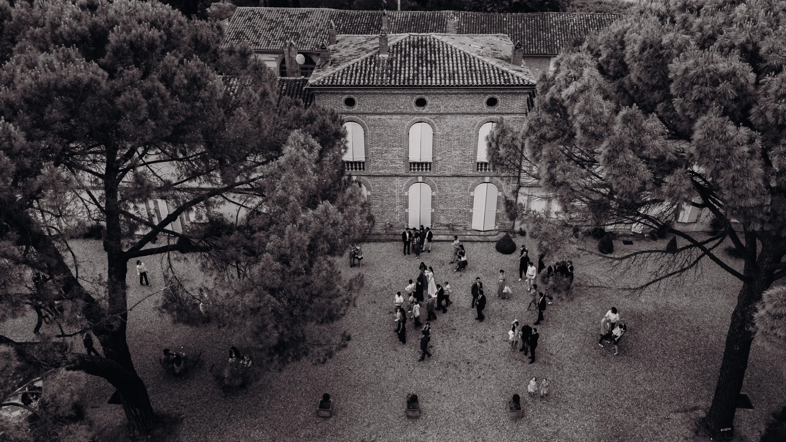 Une scène en noir et blanc vue de drone au Domaine de Gailhaguet, proche Toulouse, montrant les invités d'un mariage suivre les mariés vers le vin d'honneur
