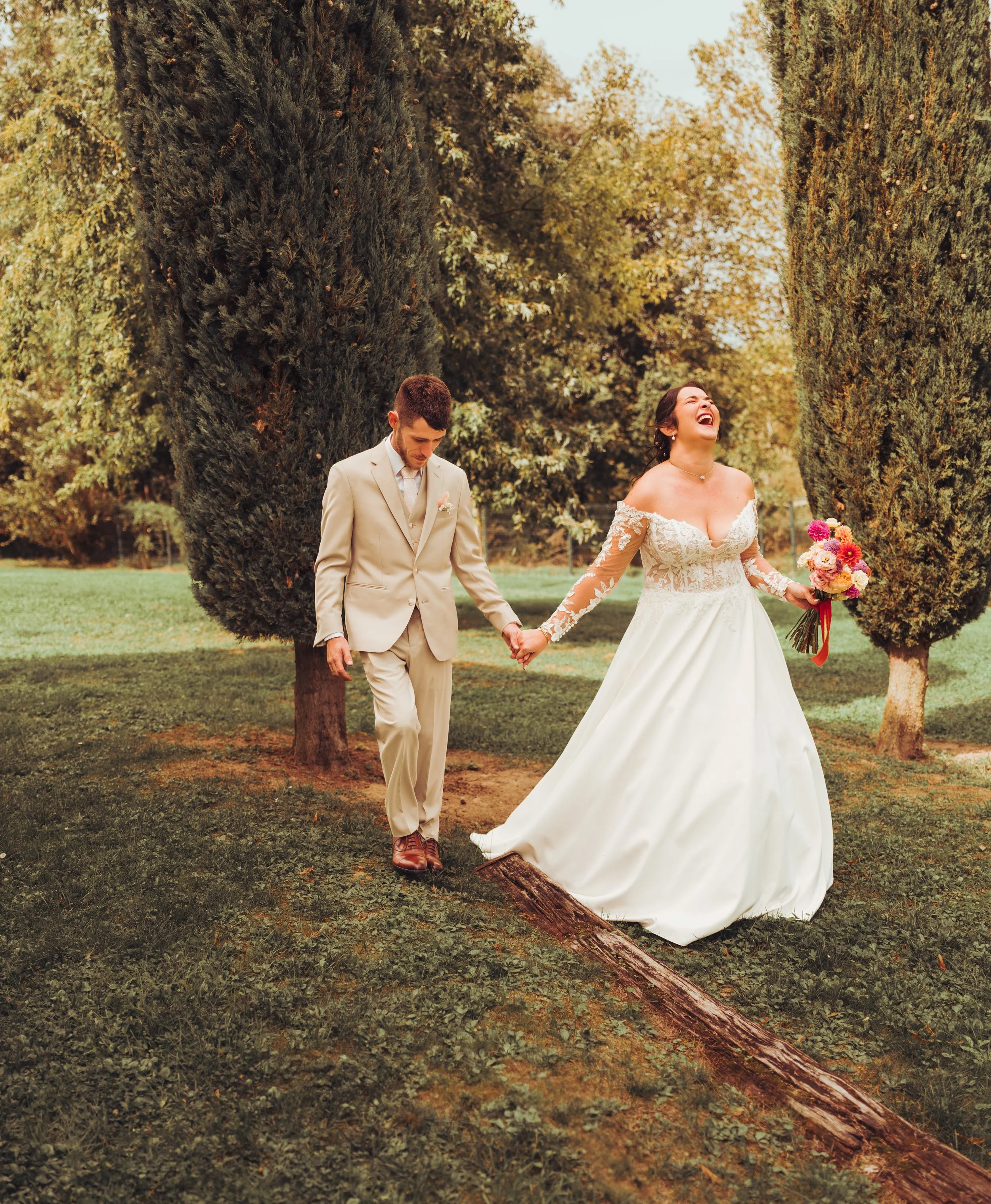 Un couple marié marche main dans la main dans le parc du Mas des Canelles proche de Toulouse, la femme rit en tenant un bouquet de fleurs.