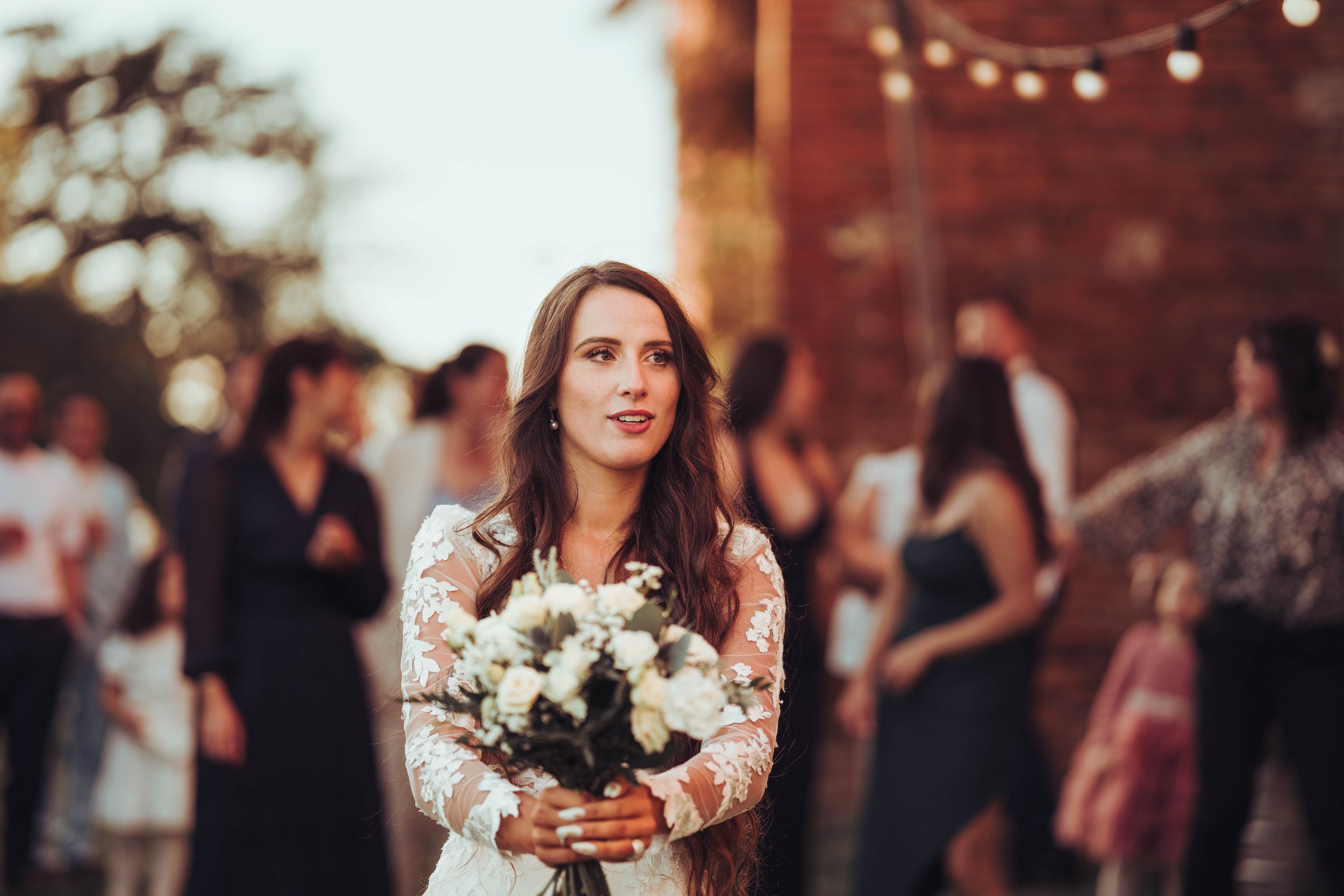 Une jeune femme en robe de mariée tient un bouquet de fleurs lors d'un mariage en extérieur avec d'autres personnes floues en arrière-plan.